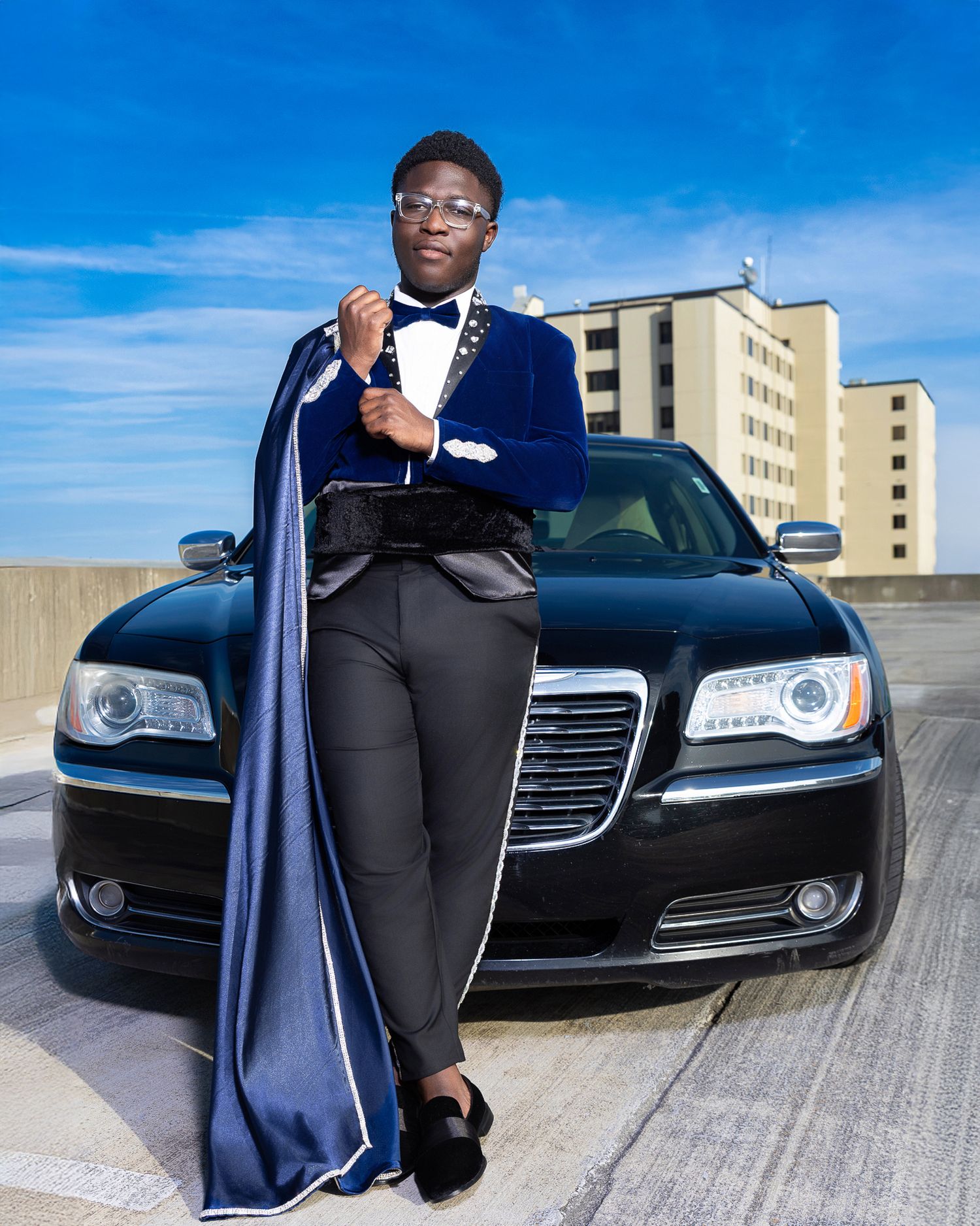Stylish young man in navy velvet tuxedo with cape poses beside a black luxury sedan outdoors.