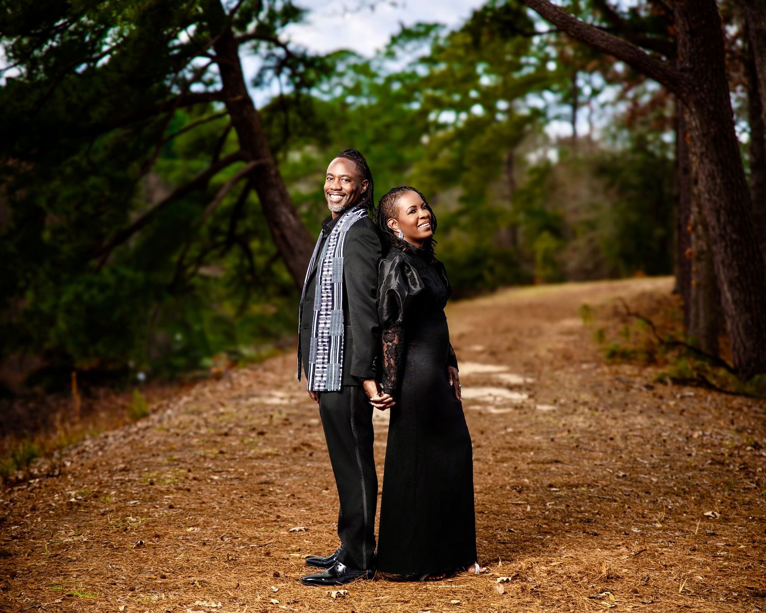 A couple poses back-to-back on a dirt path surrounded by tall trees in a serene outdoor setting.