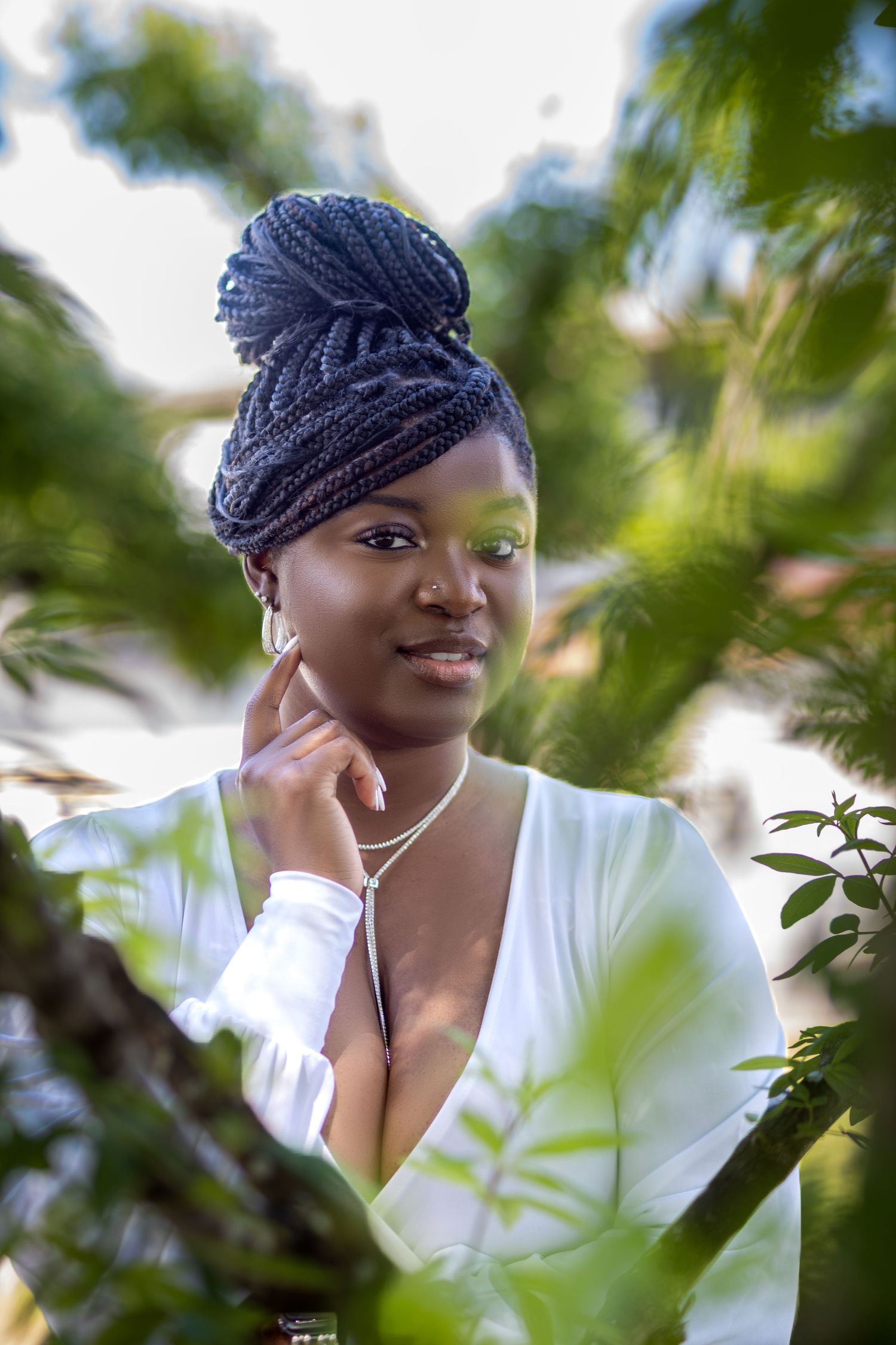 Woman with braided updo in white top poses gracefully among tropical green leaves.