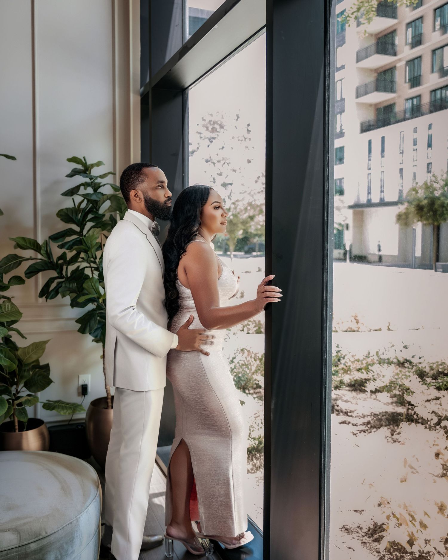 Couple in elegant white outfits embracing beside a large window
