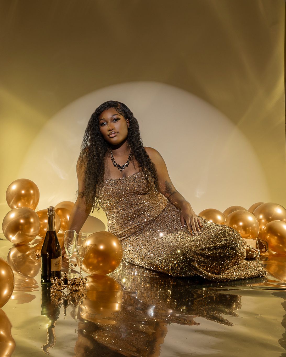 Woman in a sparkling gold gown seated among gold balloons beside champagne