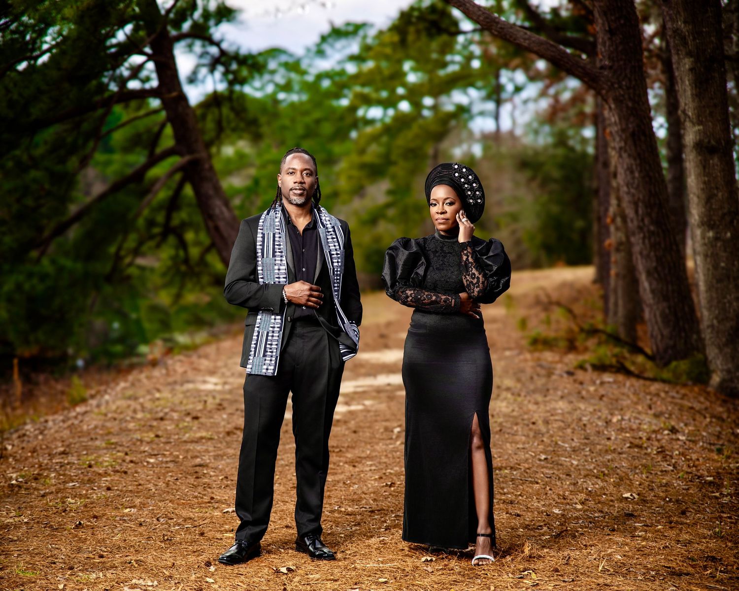 Elegant couple in matching black outfits posing on a forest path, woman in slit dress with headwrap, man in scarf.