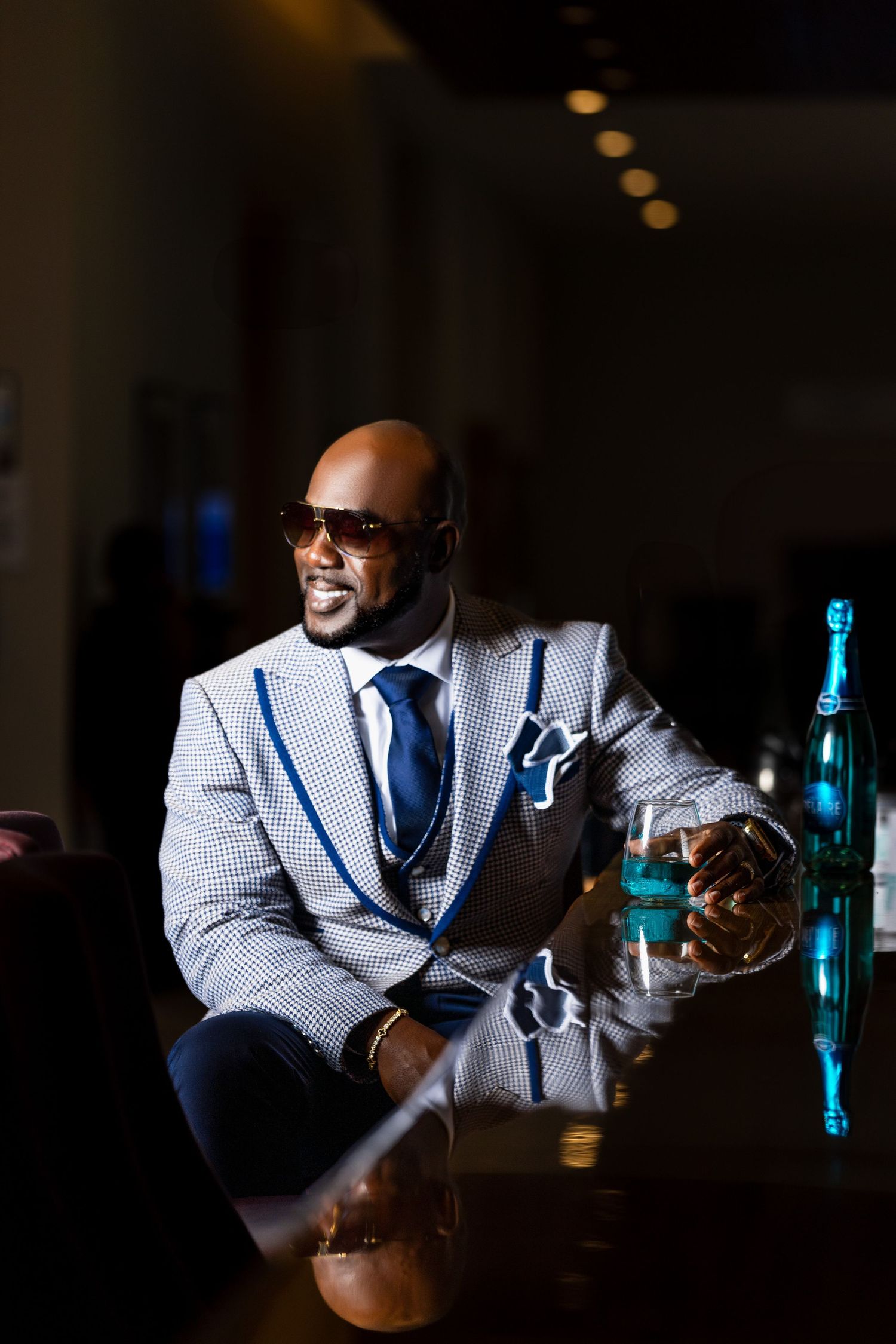 Man in a patterned suit and sunglasses seated at a table with a drink