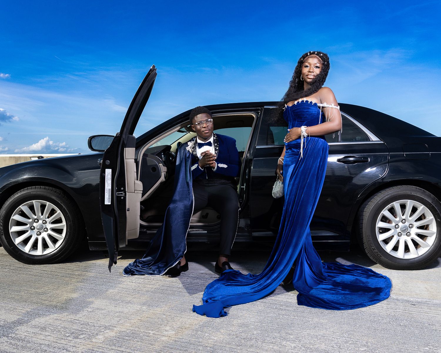 Young couple in coordinating royal blue formal wear pose elegantly beside a black luxury sedan.
