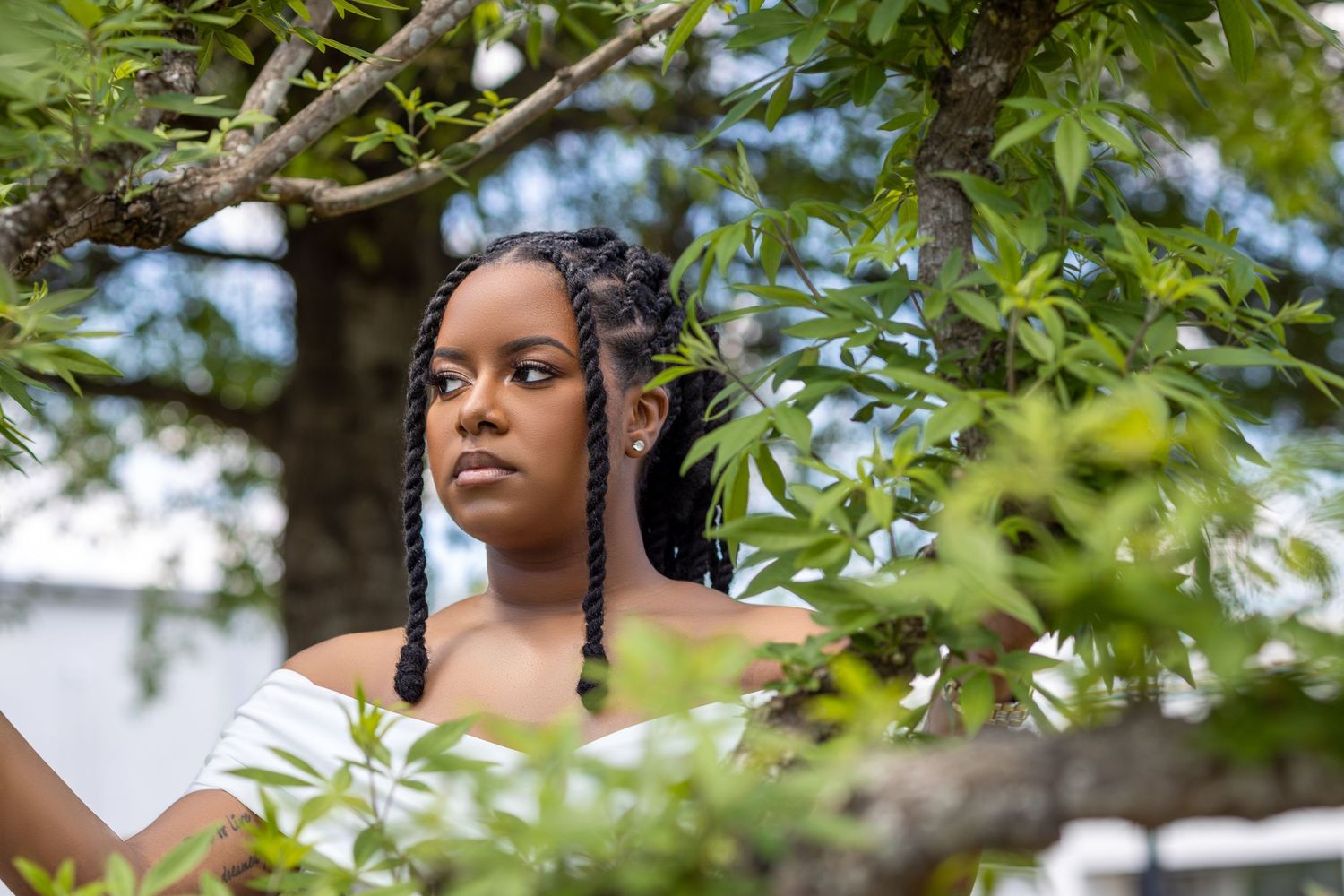 Young woman with braided hair poses among lush green tree branches outdoors.
