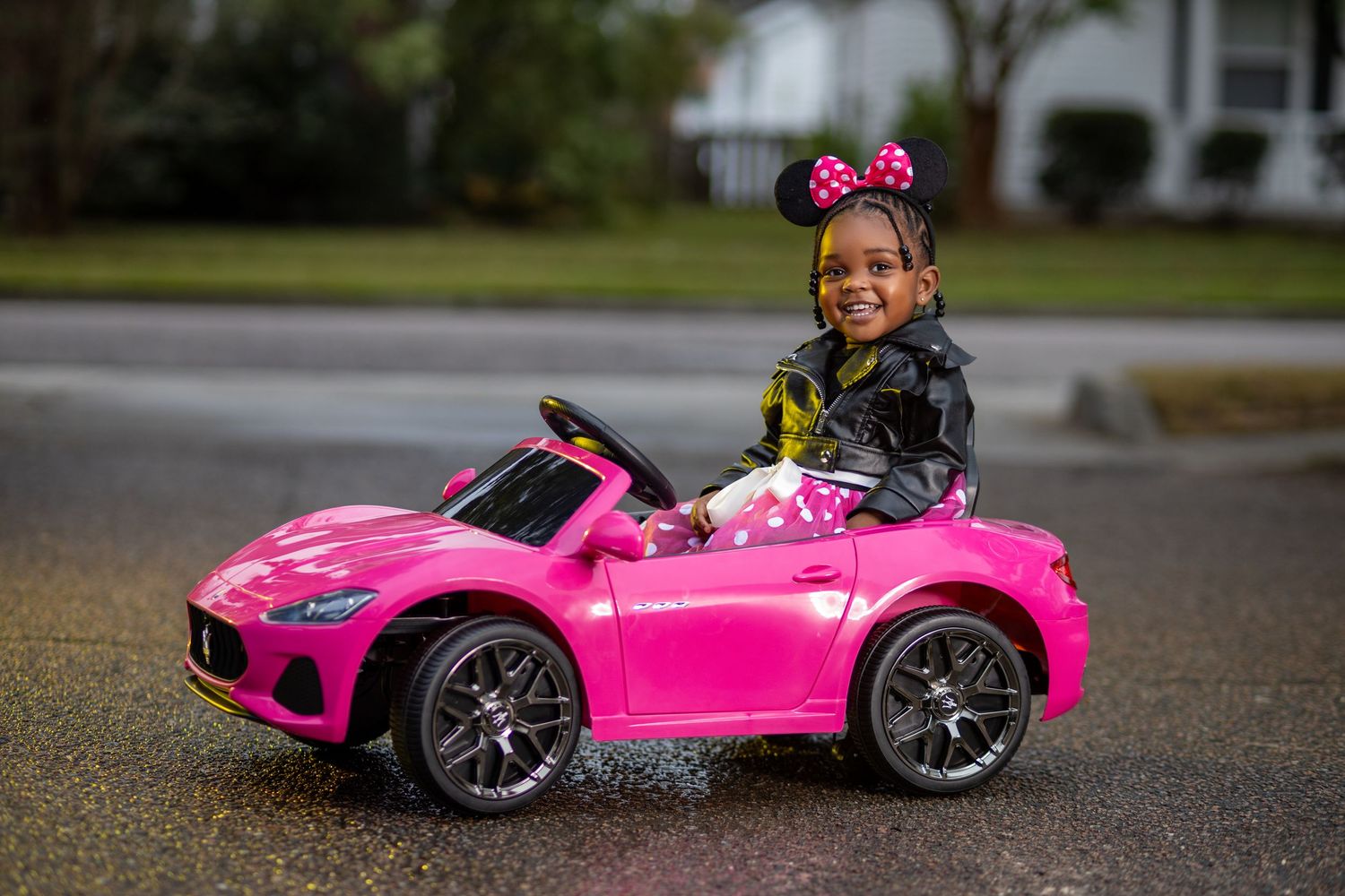 Smiling girl in Minnie Mouse costume drives pink Maserati toy car on street.