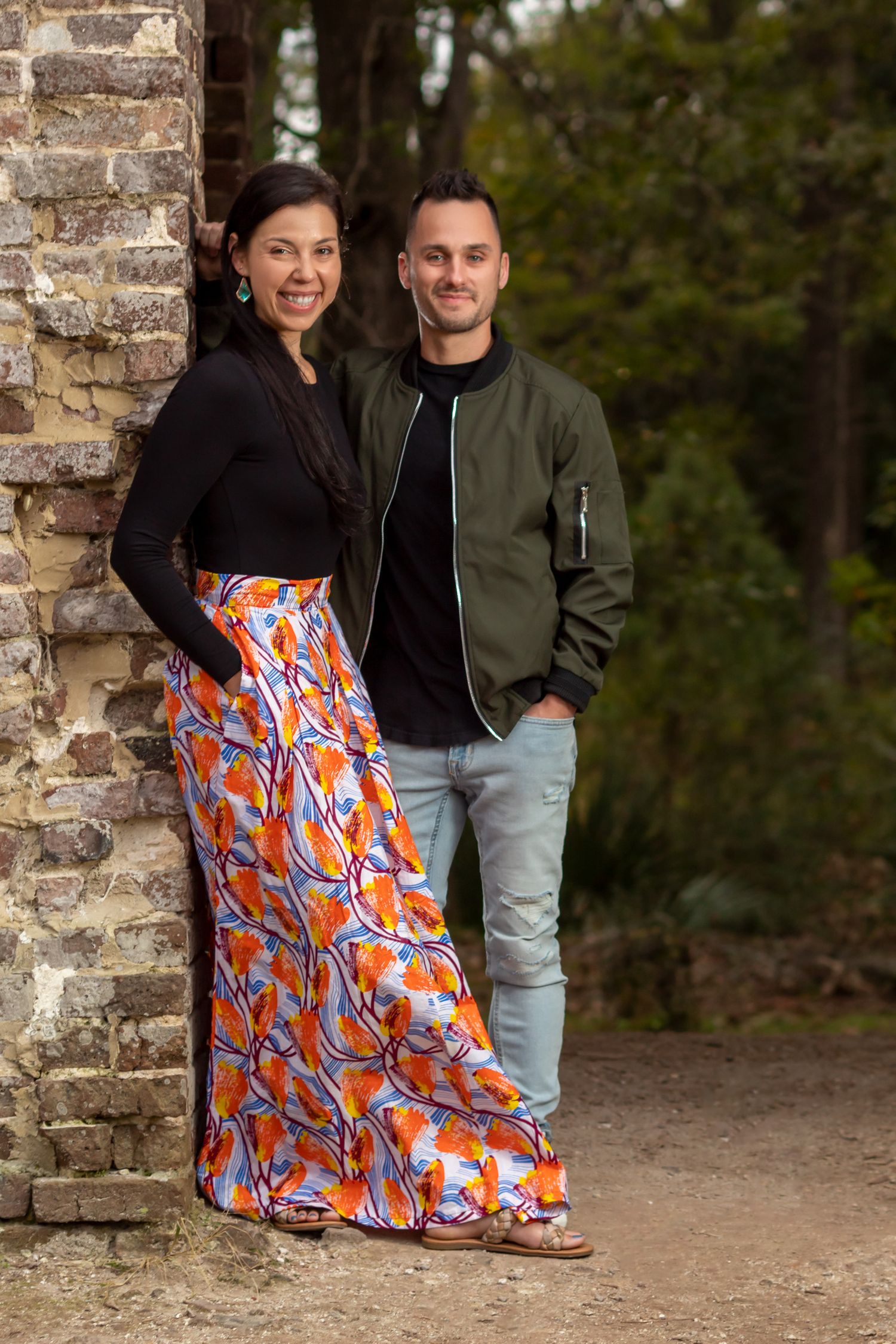 Couple posing outdoors near stone wall, woman in colorful maxi skirt, man in green bomber jacket and jeans.