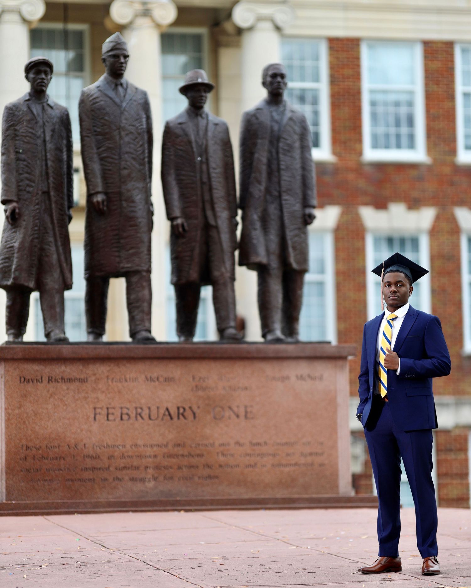 Graduate in a blue suit and cap posing beside the February One statues