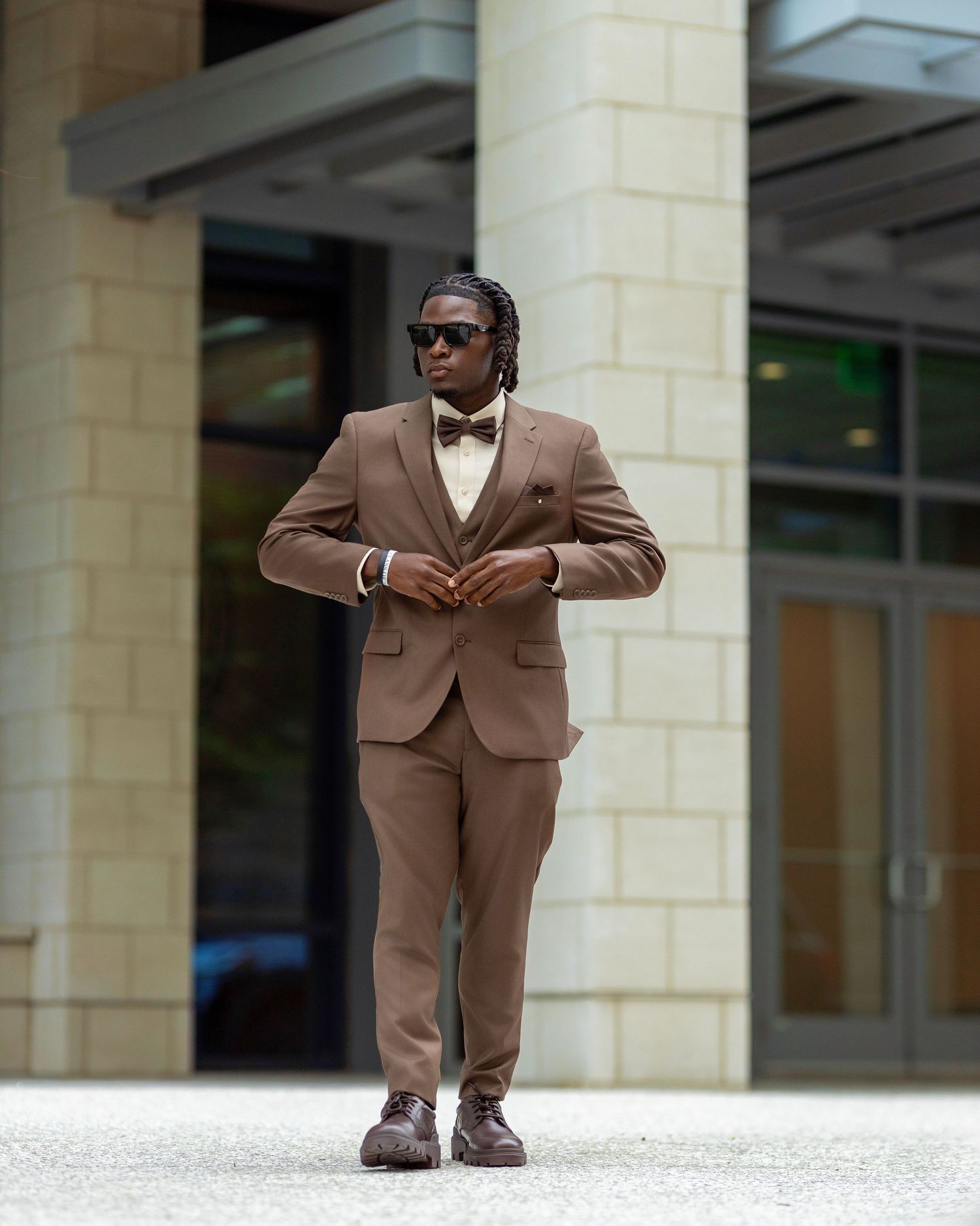 Confident man in brown three-piece suit and sunglasses standing outside modern building.