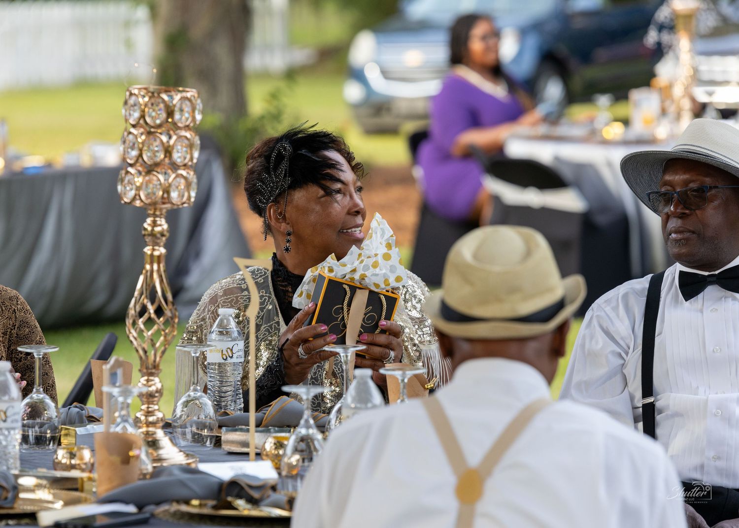 Woman in sequined outfit reads card at glamorous outdoor evening dinner celebration event.