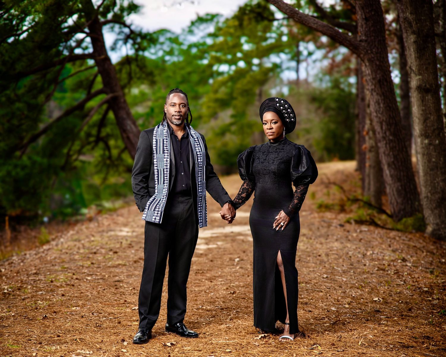Couple holding hands on a wooded path, man in black suit with patterned scarf, woman in black dress with puff sleeves.