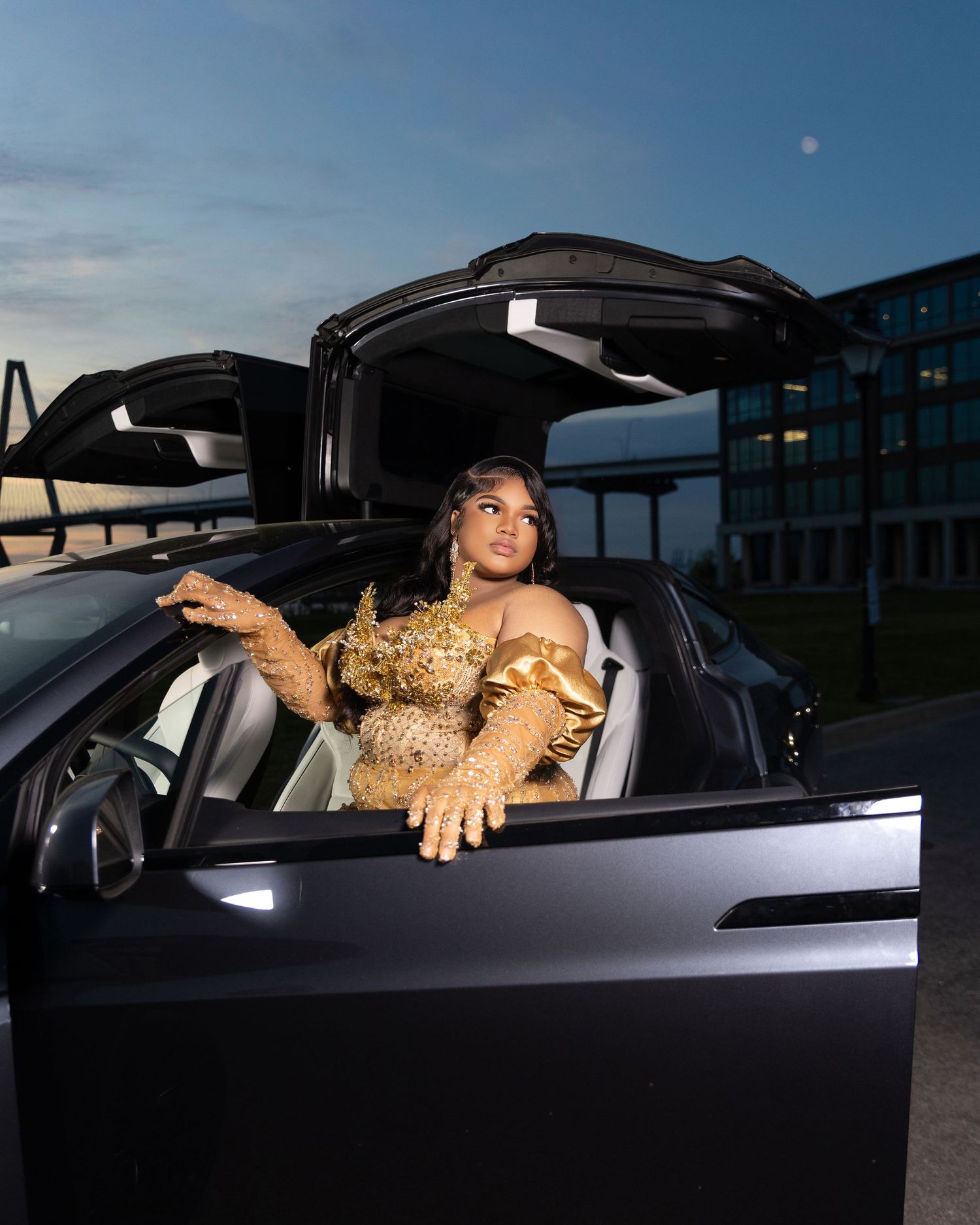 Woman in gold gown poses in Tesla Model X with gull-wing doors open at dusk.