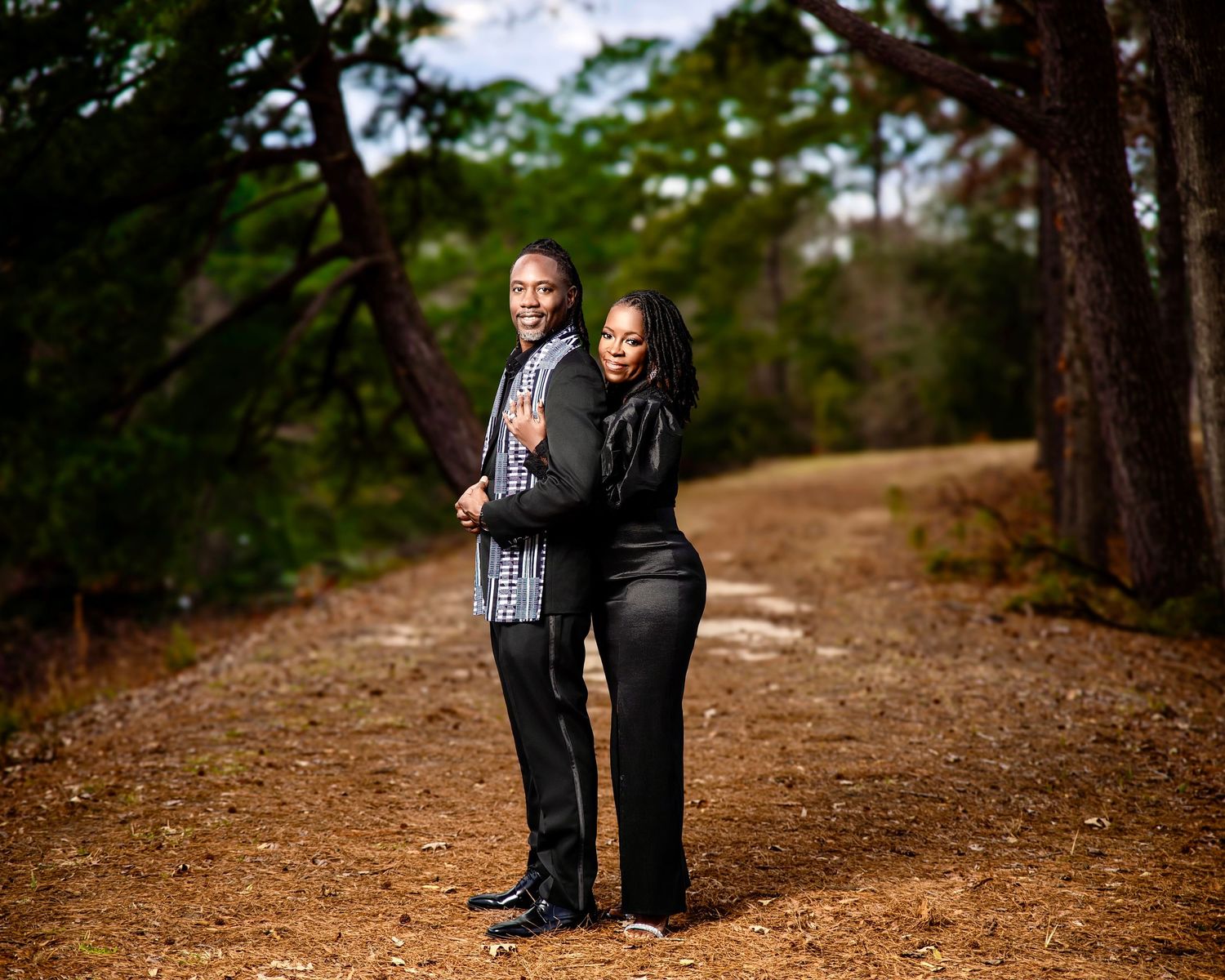 Couple posing on a dirt path surrounded by tall trees, man in plaid shirt and woman in black outfit.