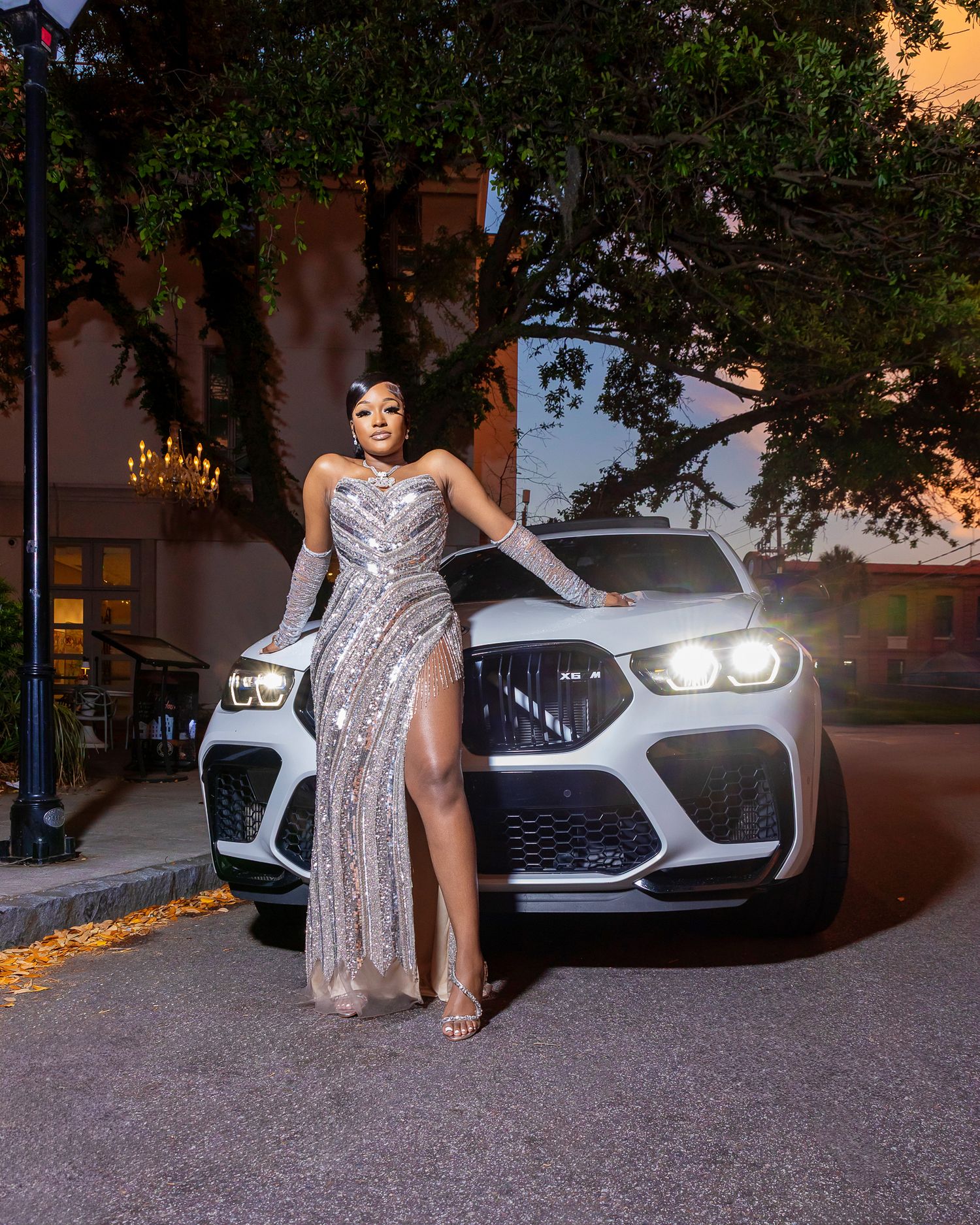 Elegant woman in silver sequined gown with thigh slit posing in front of white BMW SUV.