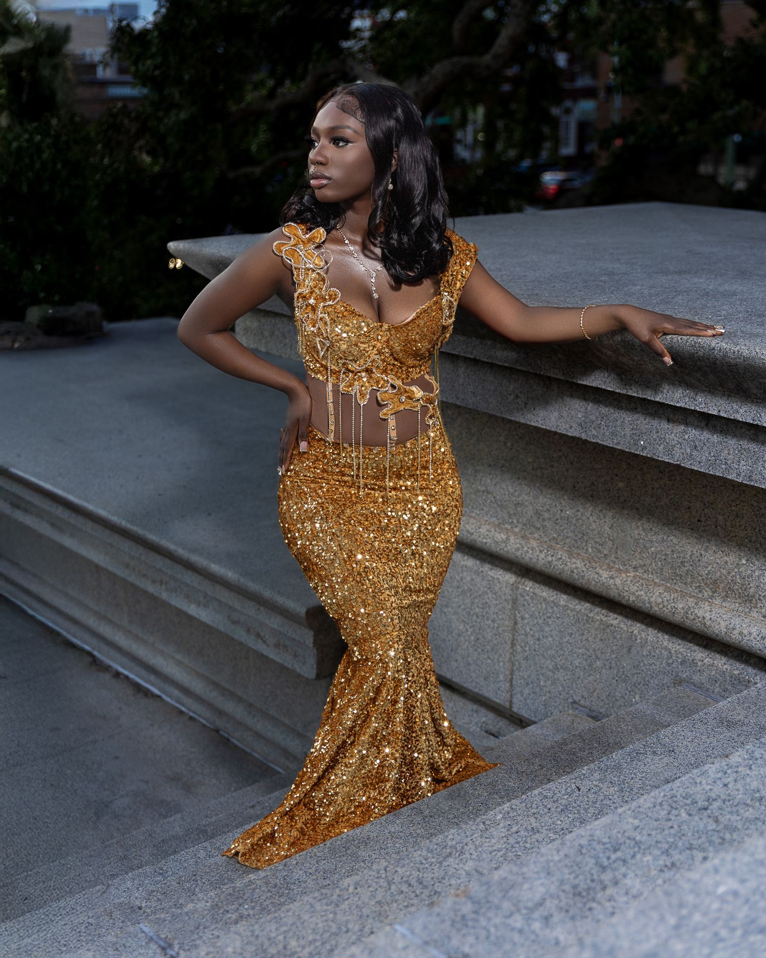 Elegant woman in gold sequin mermaid dress strikes a pose on outdoor stone steps at dusk.