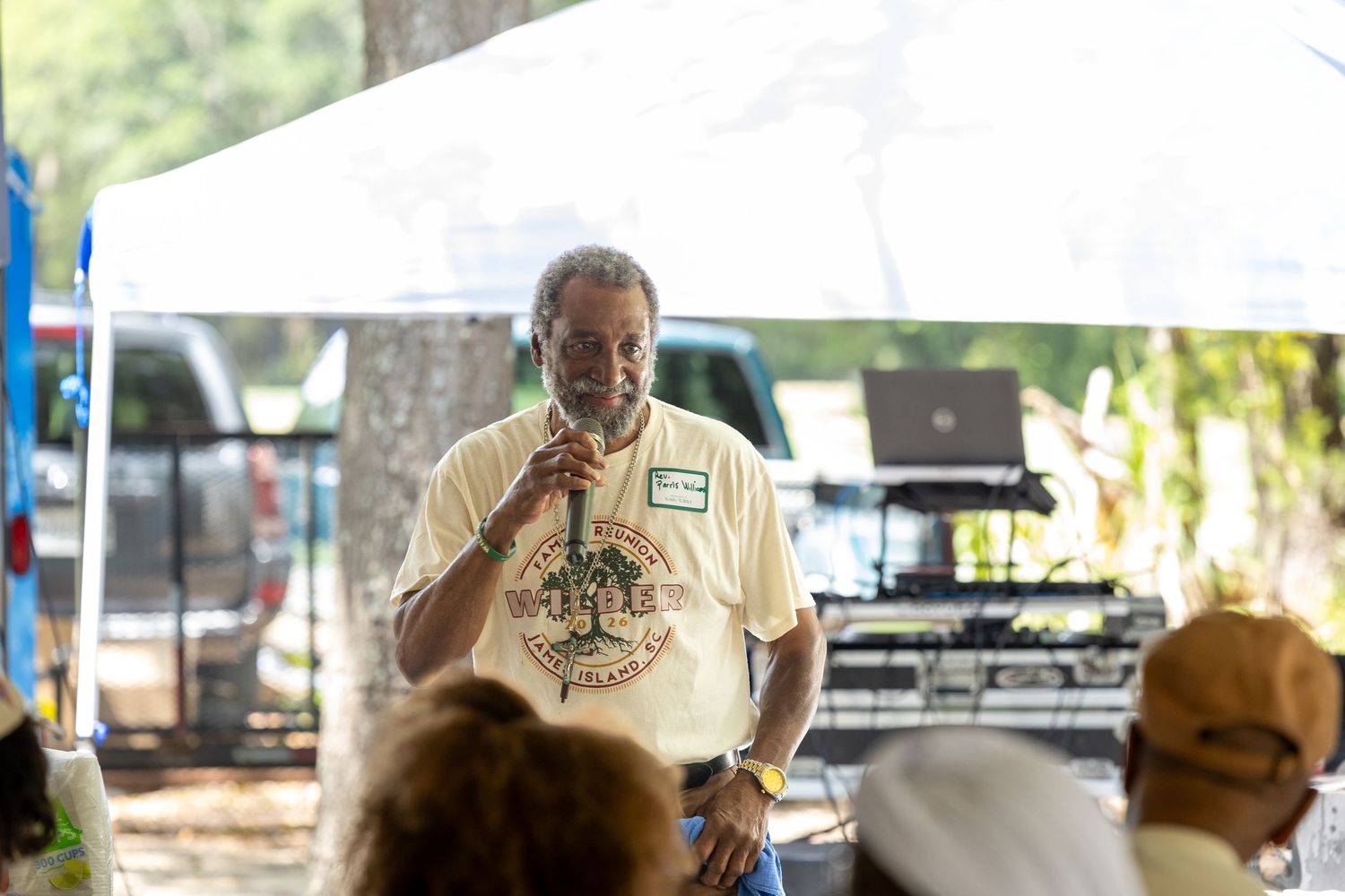 Man in family reunion t-shirt speaks at microphone under white tent at outdoor gathering.