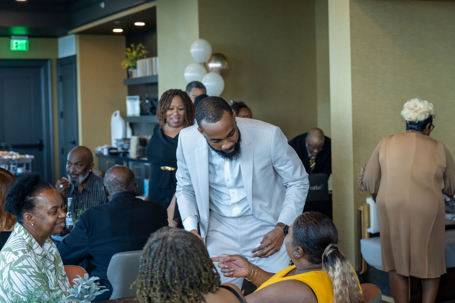 Man in white outfit leans over to greet seated guests at an indoor celebration.