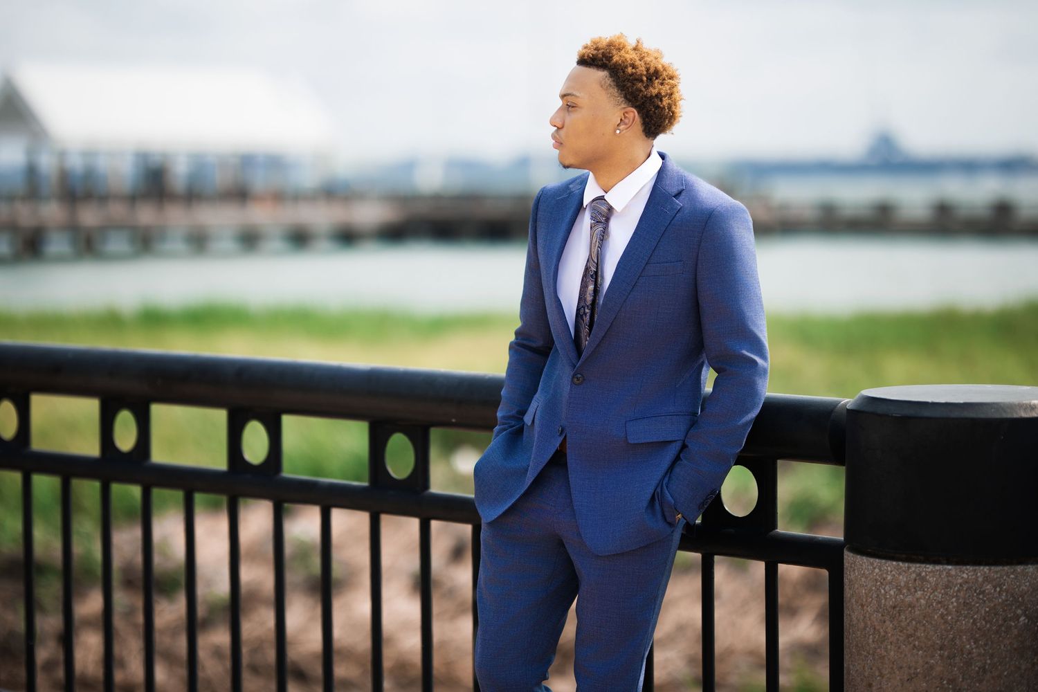Young man in blue suit leans on railing with waterfront view behind him.