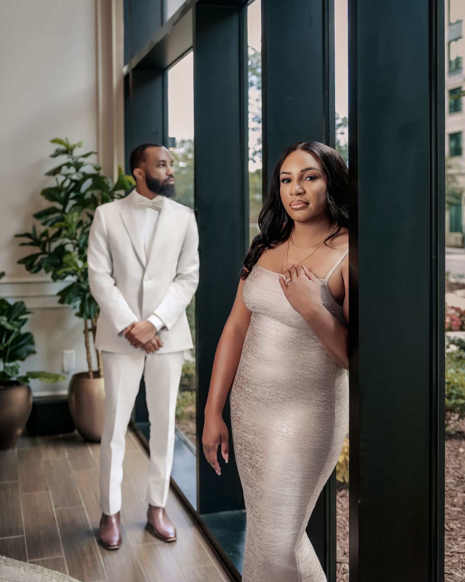 Couple in elegant white outfits embracing beside a large window