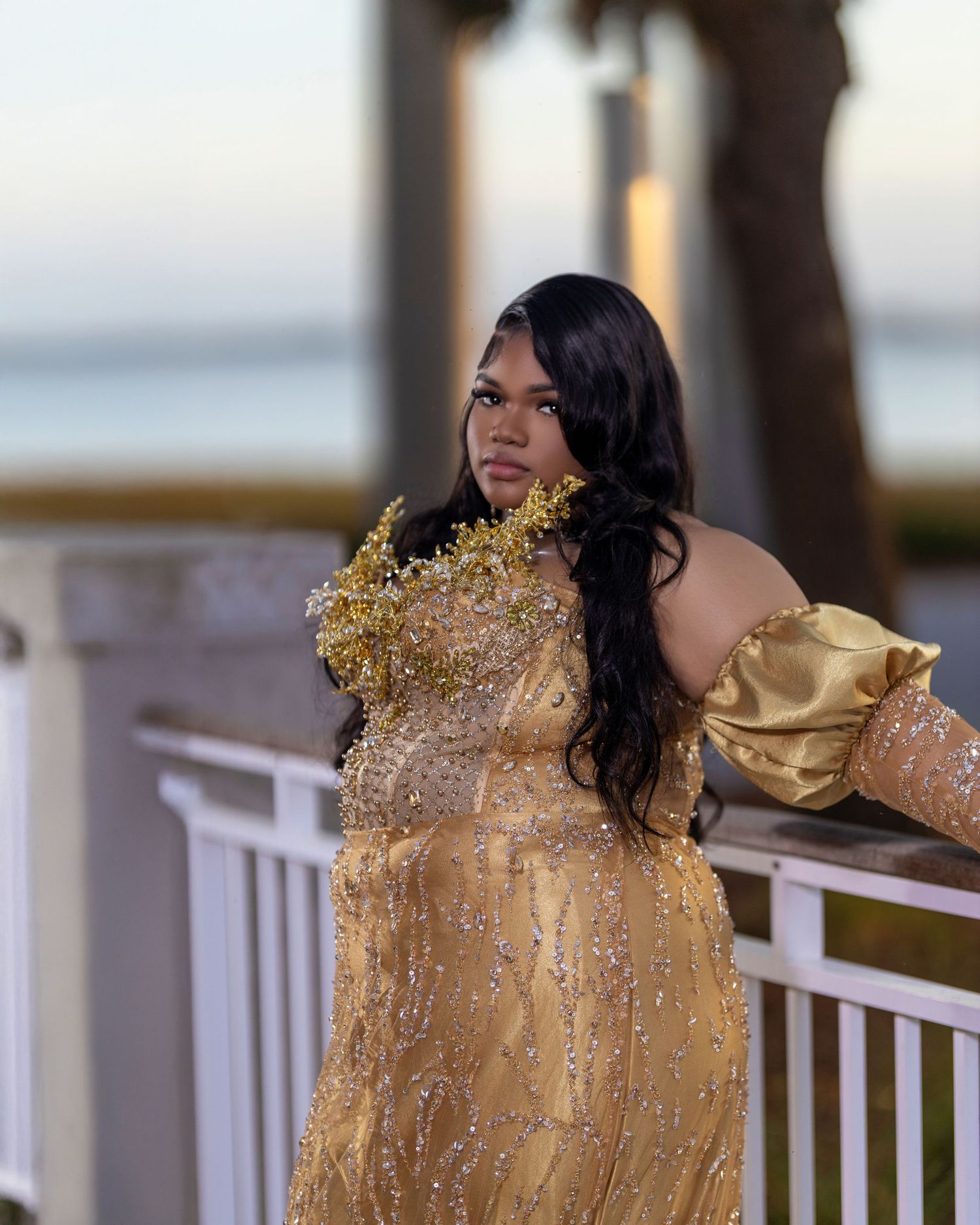 Woman in gold beaded off-shoulder gown with floral detail posing on seaside balcony.