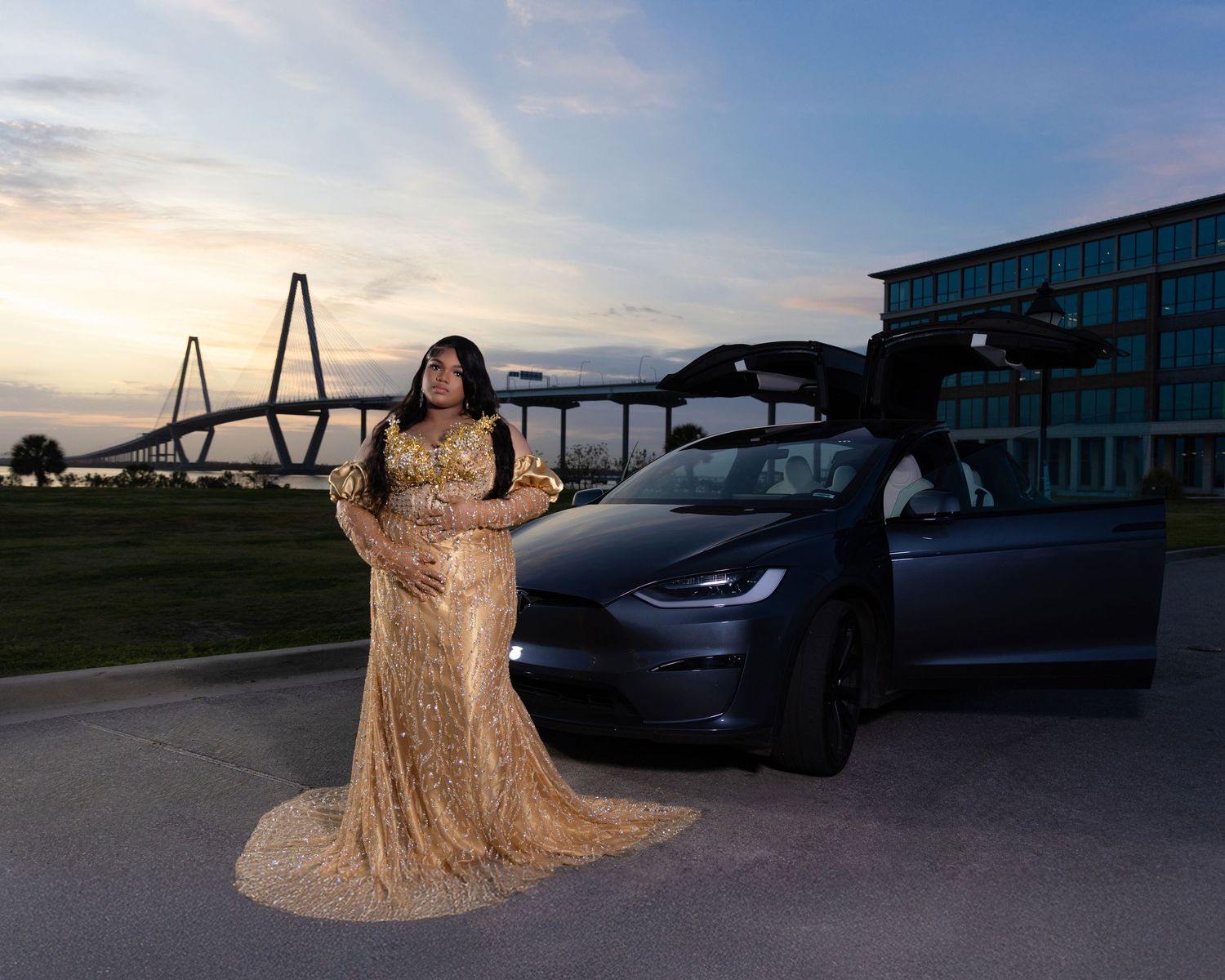 Woman in gold sequin gown stands beside Tesla Model X near bridge at sunset.