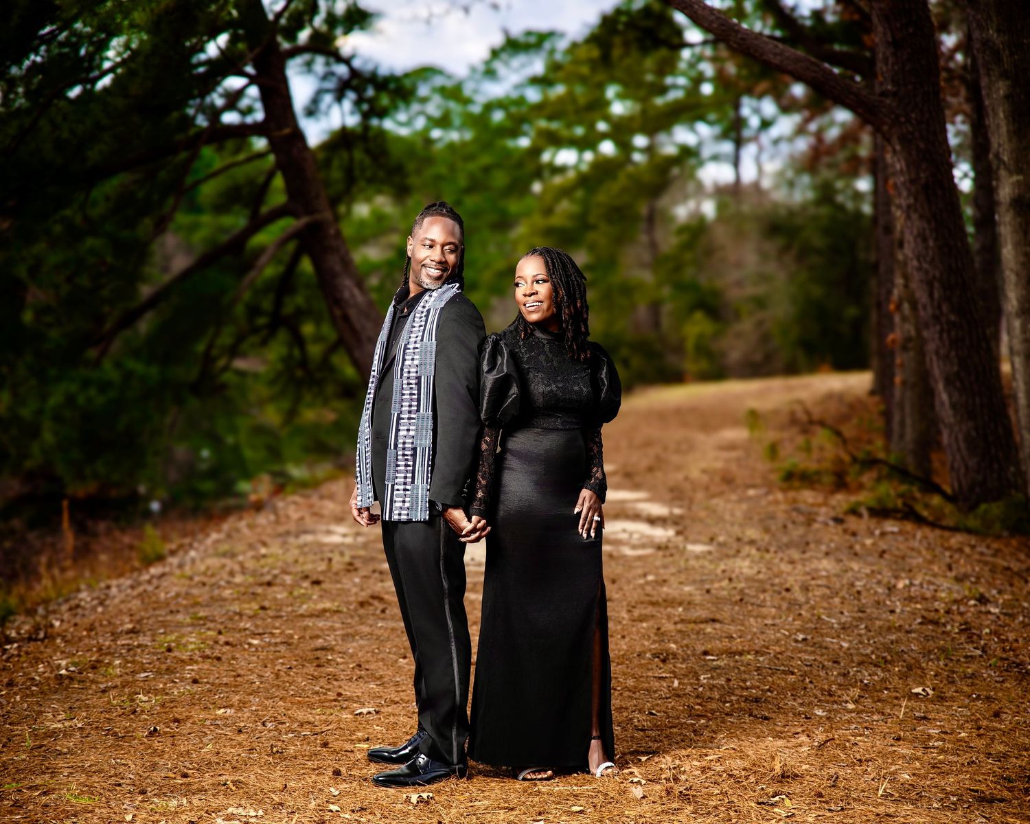 Couple holding hands on a forest path, man in gray blazer, woman in elegant black gown, surrounded by tall pine trees.