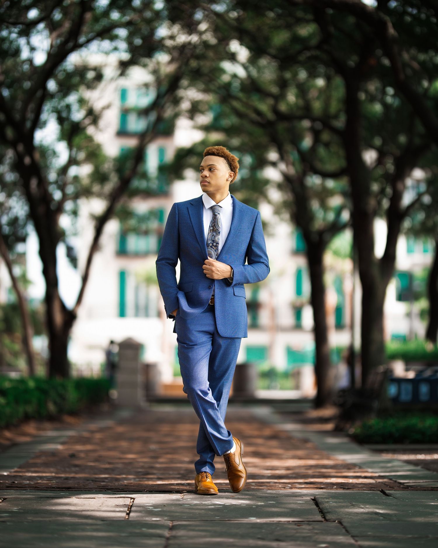 Man in blue suit and tie walking confidently through a tree-lined park path with blurred building background.