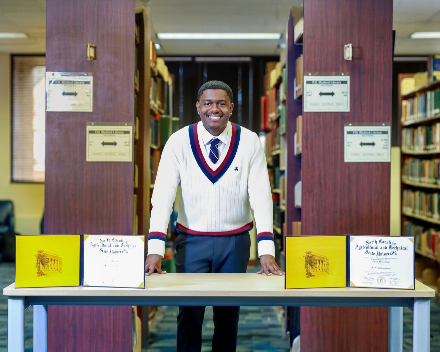 Smiling graduate standing behind two displayed diplomas in a library