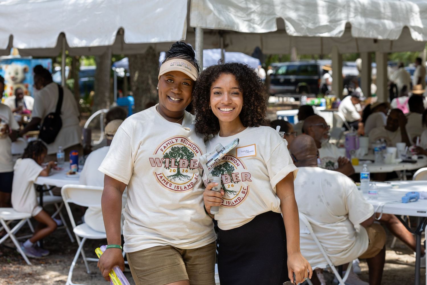 Two women smiling together wearing matching event t-shirts at an outdoor gathering.