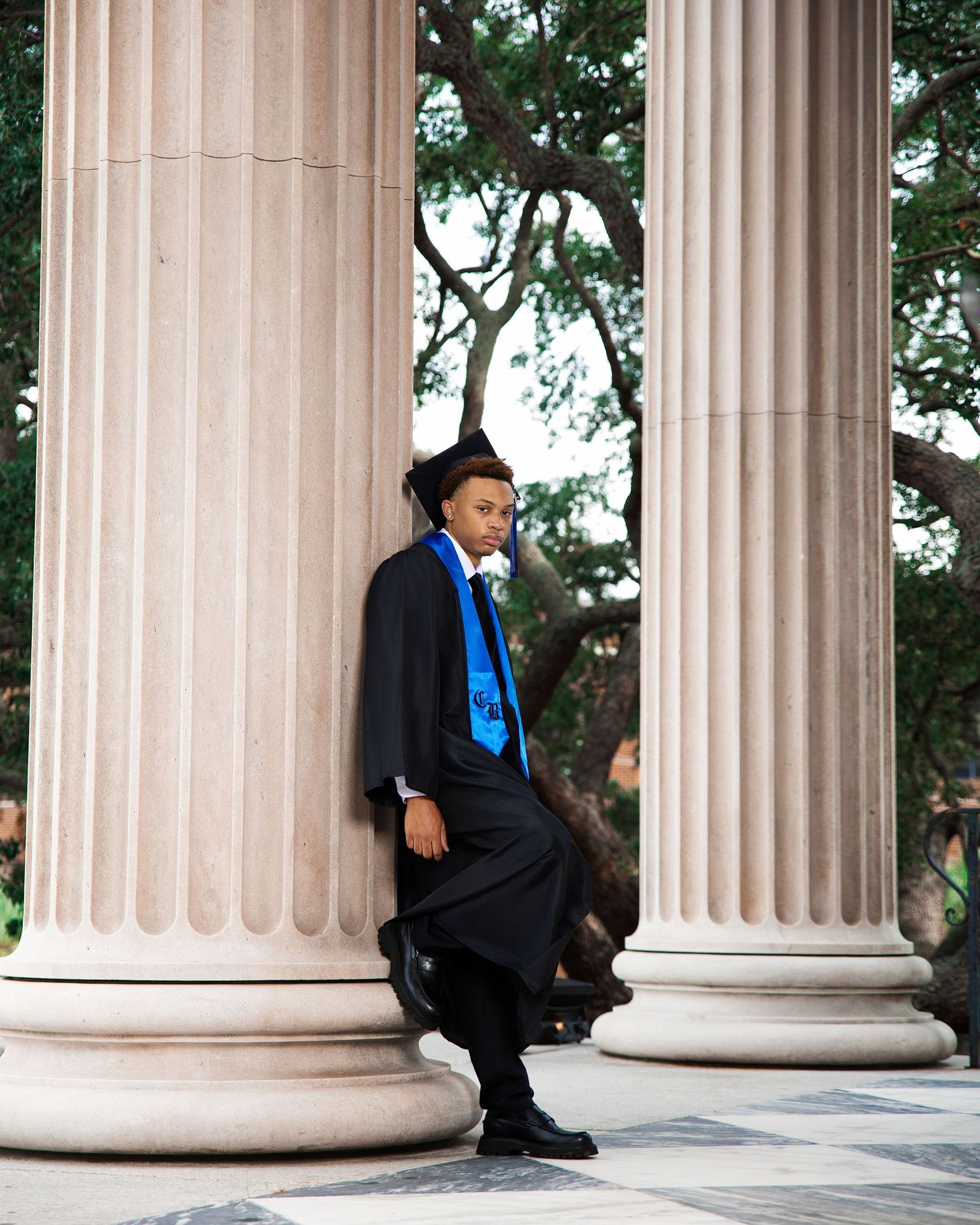 Graduate in black cap and gown with blue stole leans against classical columns outdoors.
