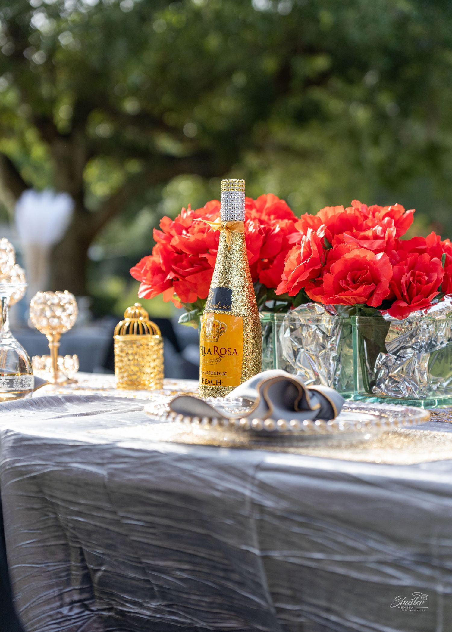 Elegant outdoor table setting with red roses, gold glitter champagne bottle, and crystal decor.