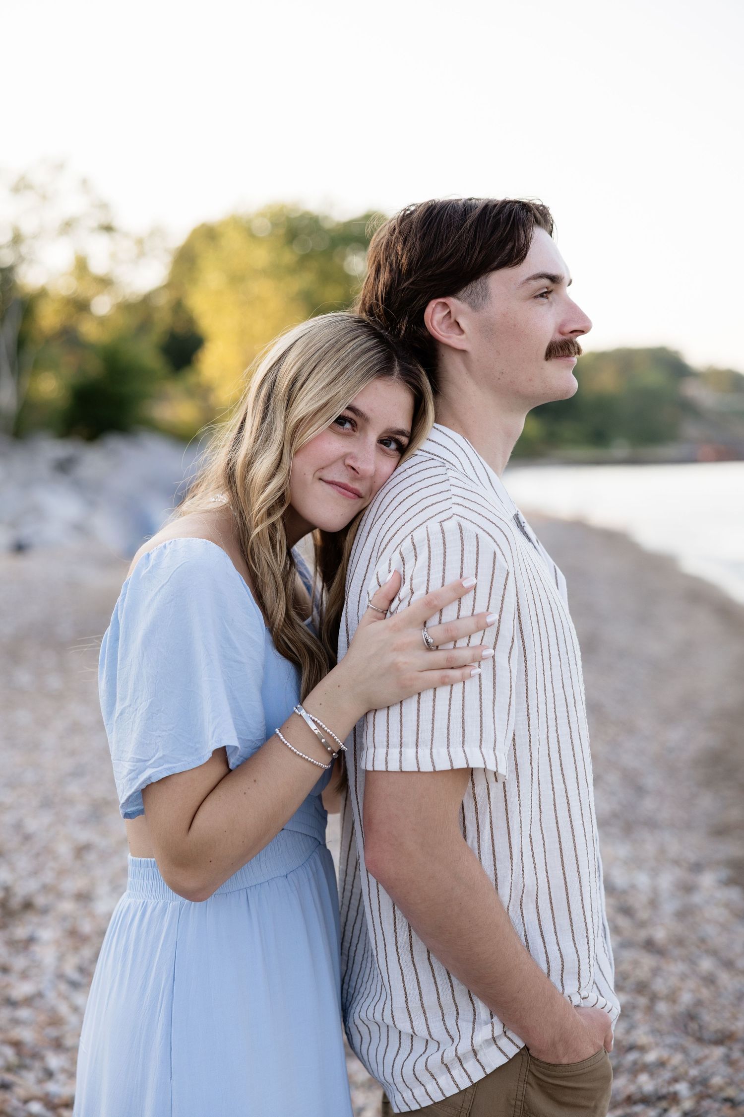 A couple poses on a rocky beach, woman in blue dress leaning on man in striped shirt at golden hour.