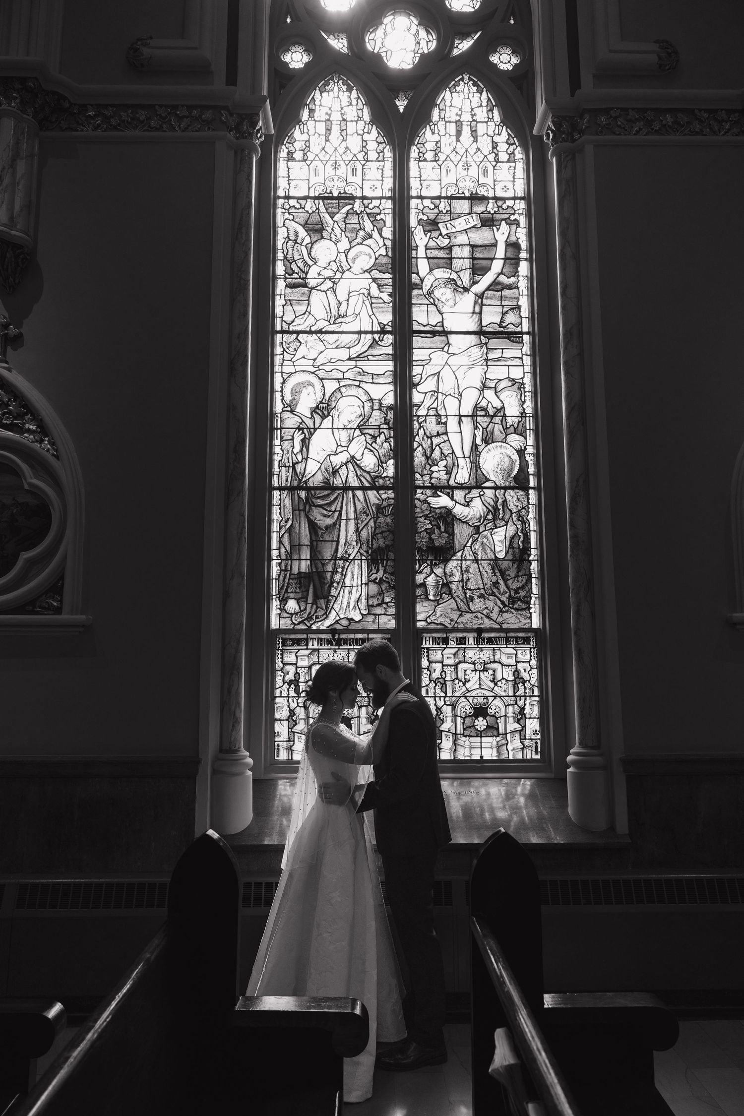 Black and white photo of a bride and groom sharing an intimate moment before a stunning stained glass church window.