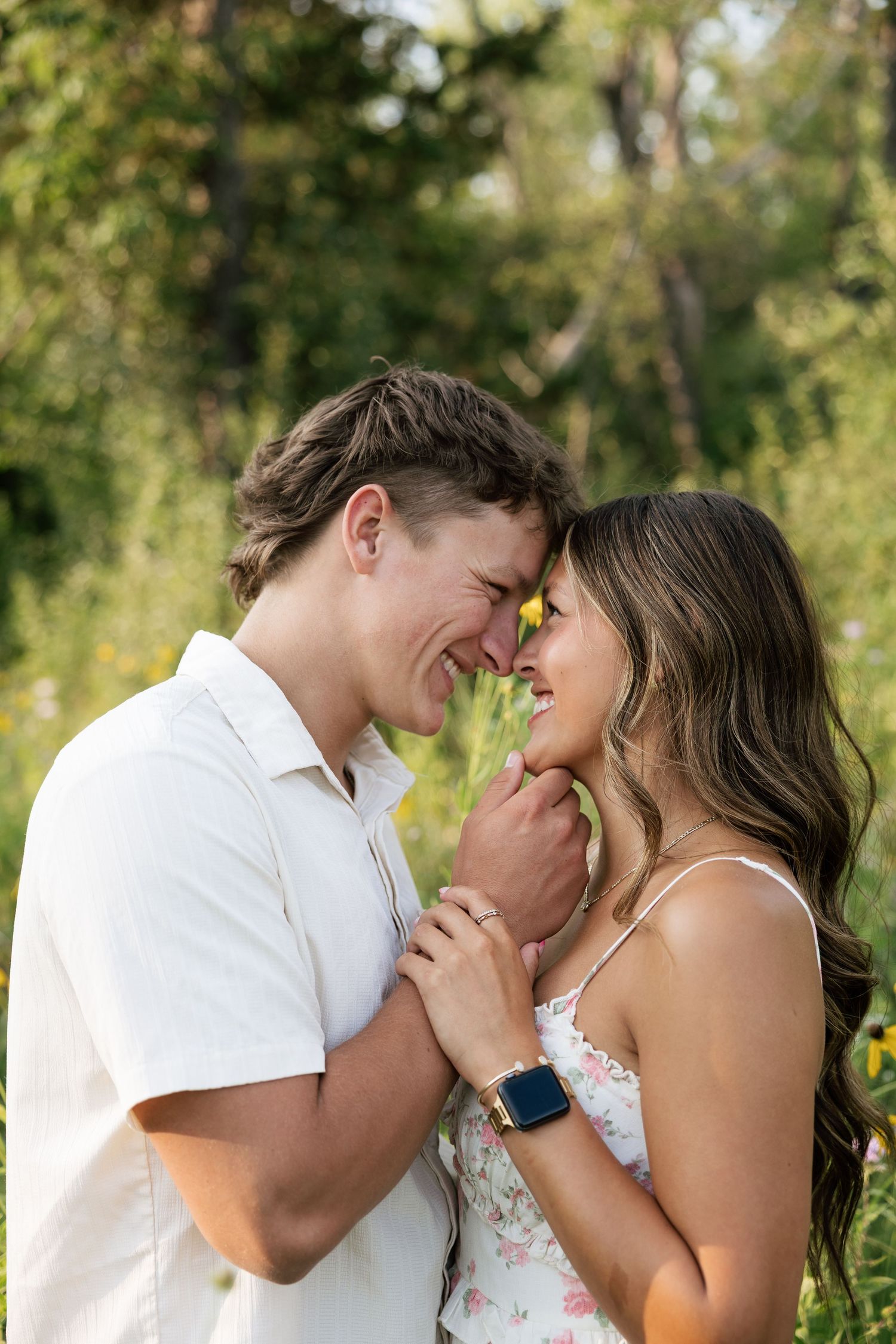 Couple smiling and touching foreheads in a sunny garden, holding hands with a floral dress and white shirt.