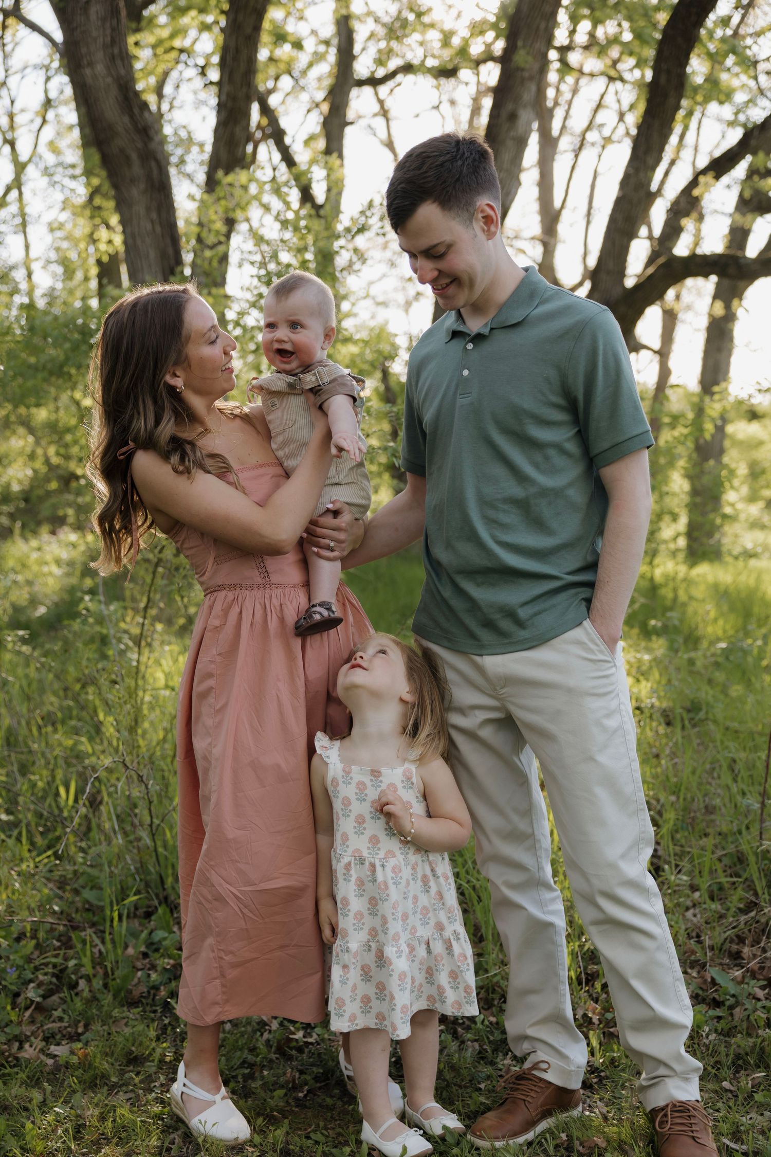 Joyful family of four laughing together in a sunlit meadow, mother lifting happy baby while toddler looks up.