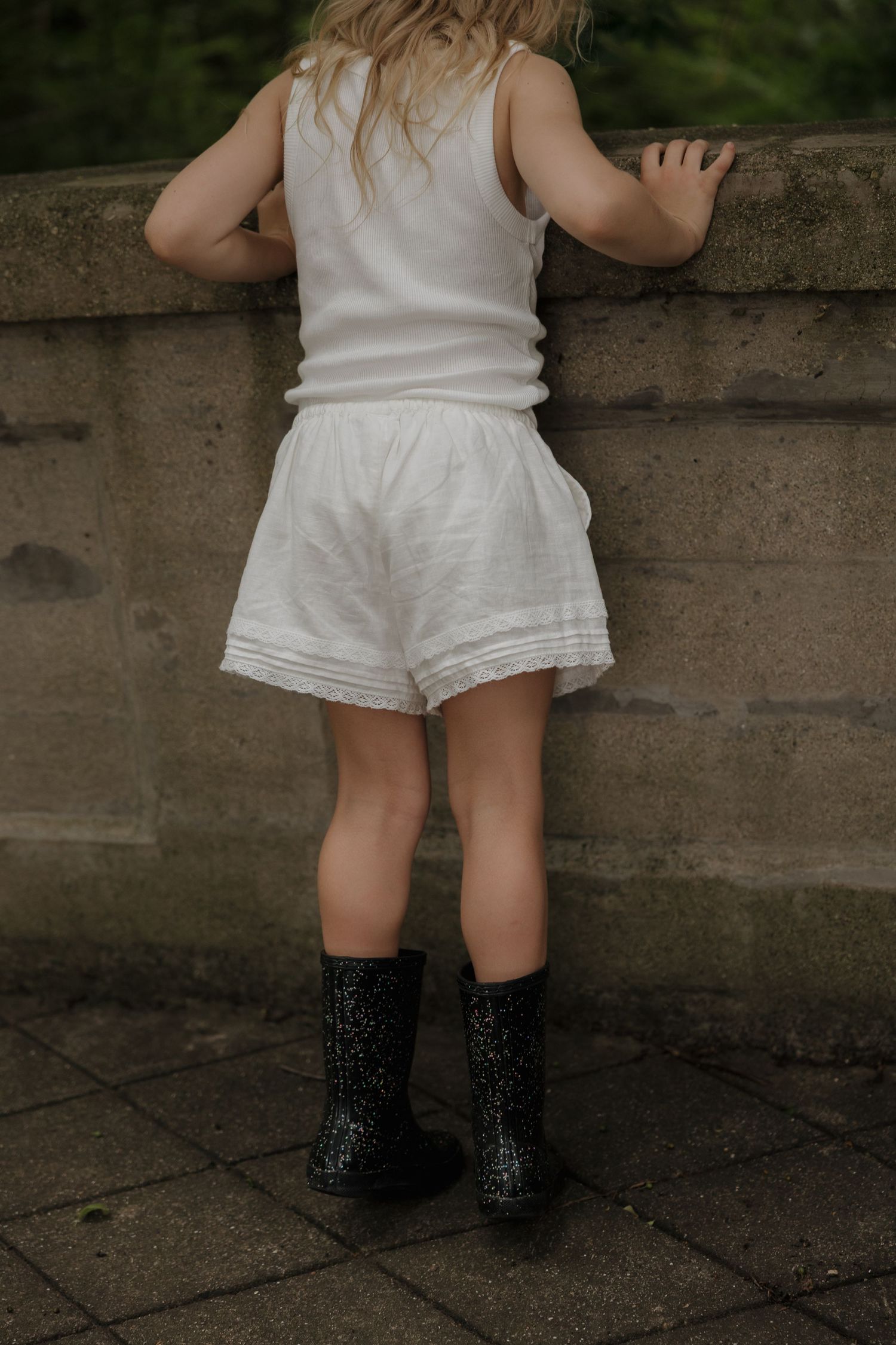 Young girl in white outfit and black glitter boots standing at stone wall outdoors.