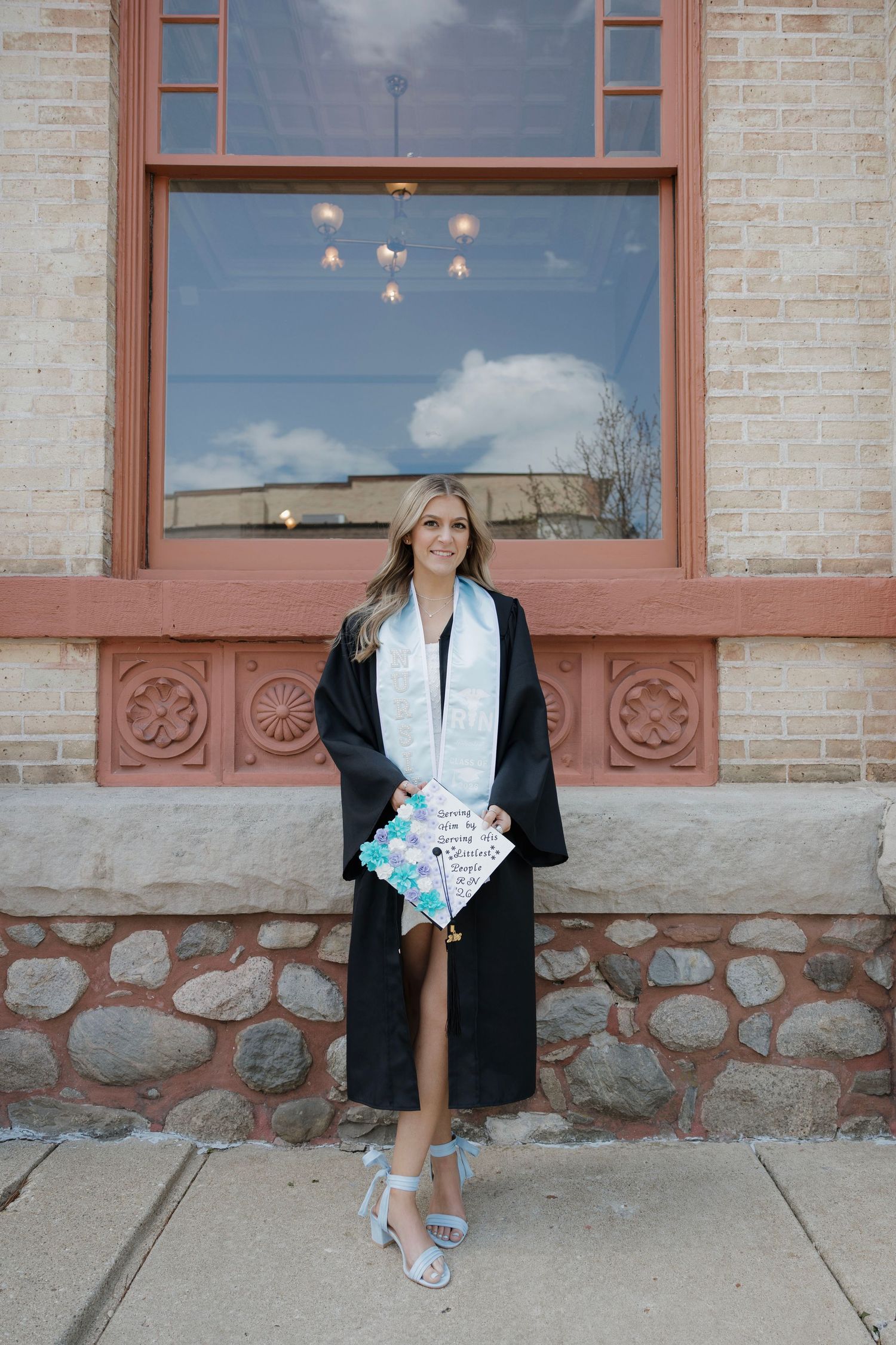 Blonde woman in graduation cap and gown holding decorated mortarboard outside brick building.
