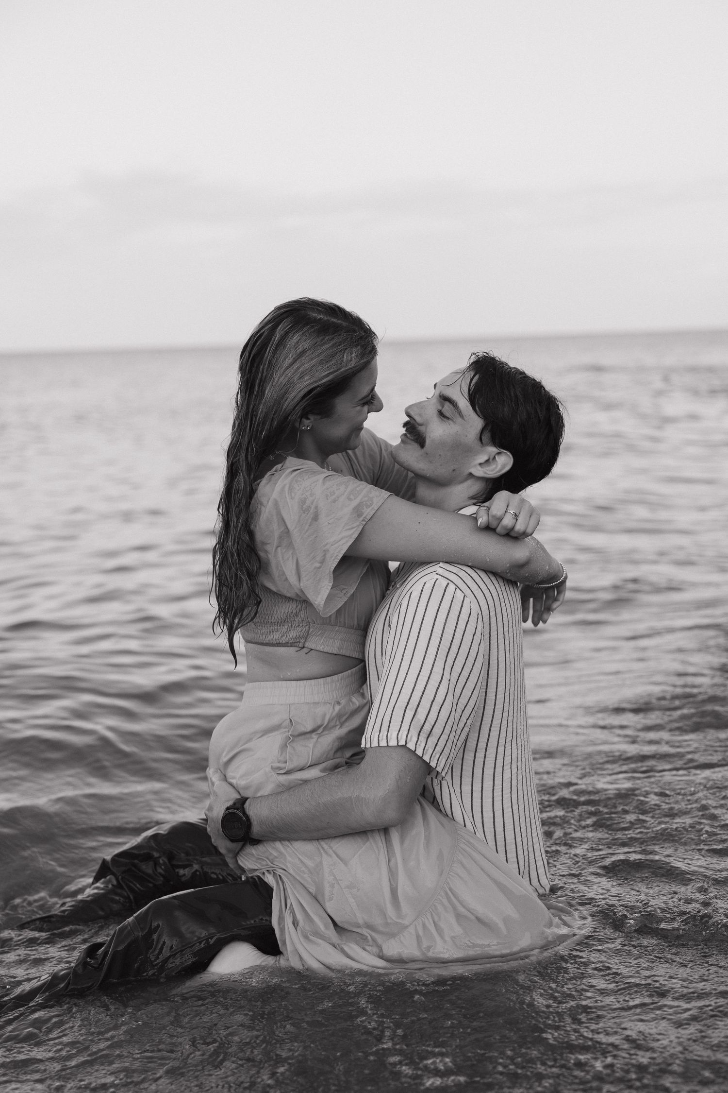 Black and white photo of a romantic couple embracing while sitting in ocean waves at dusk.