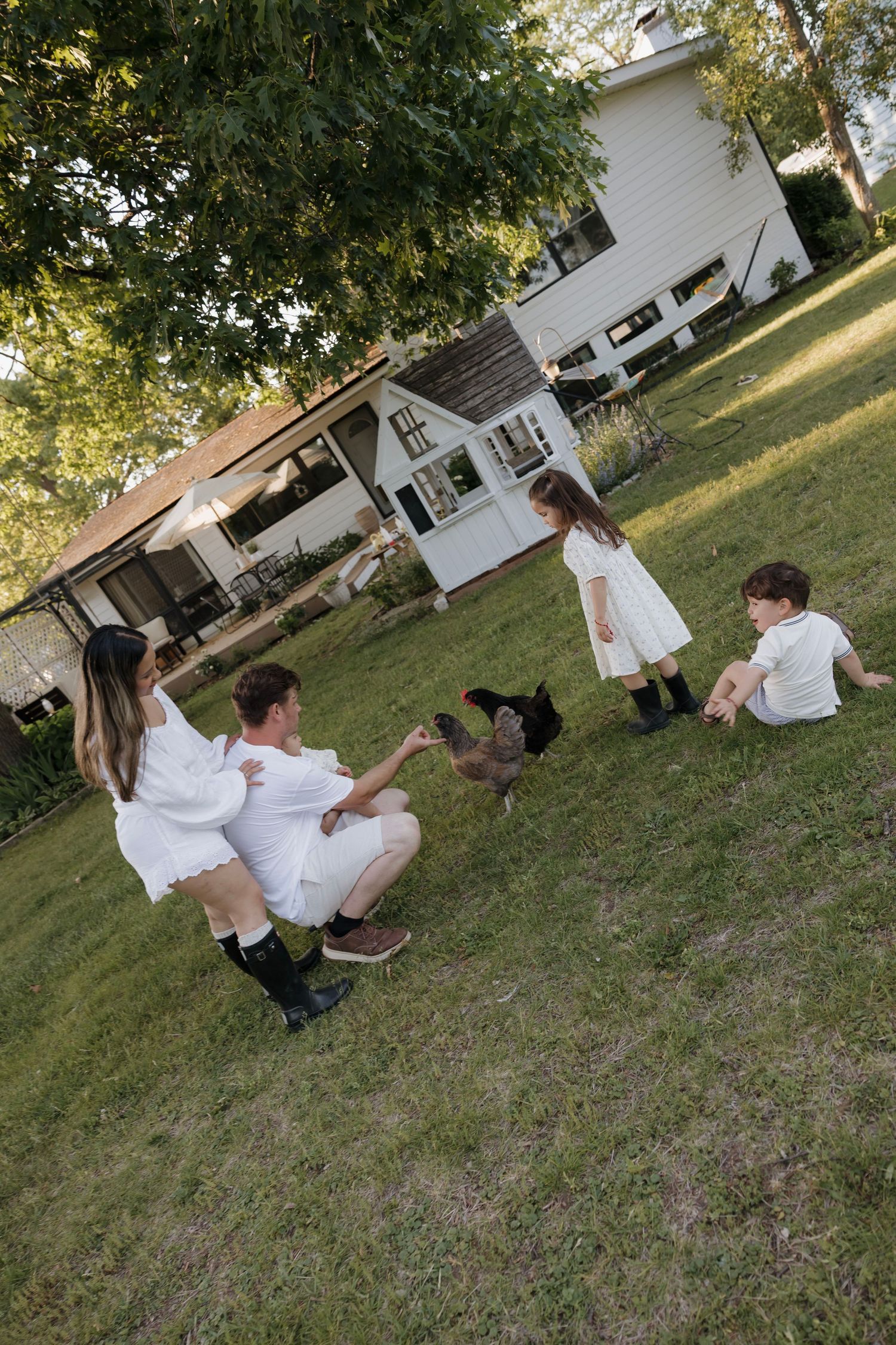 Family sitting on grass in white outfits interacting with chickens in a sunny backyard.