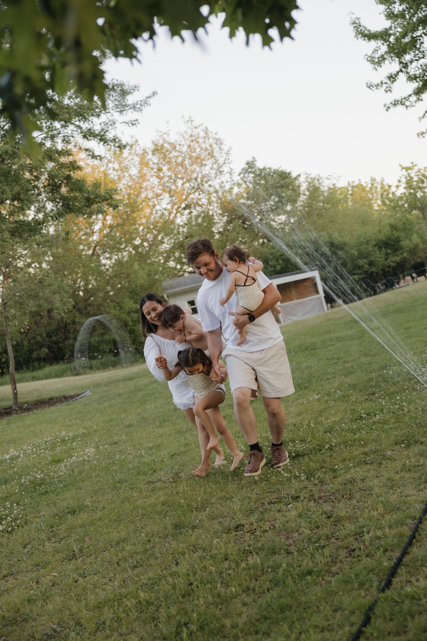 Joyful family playing together on green grass yard with sprinkler in background.