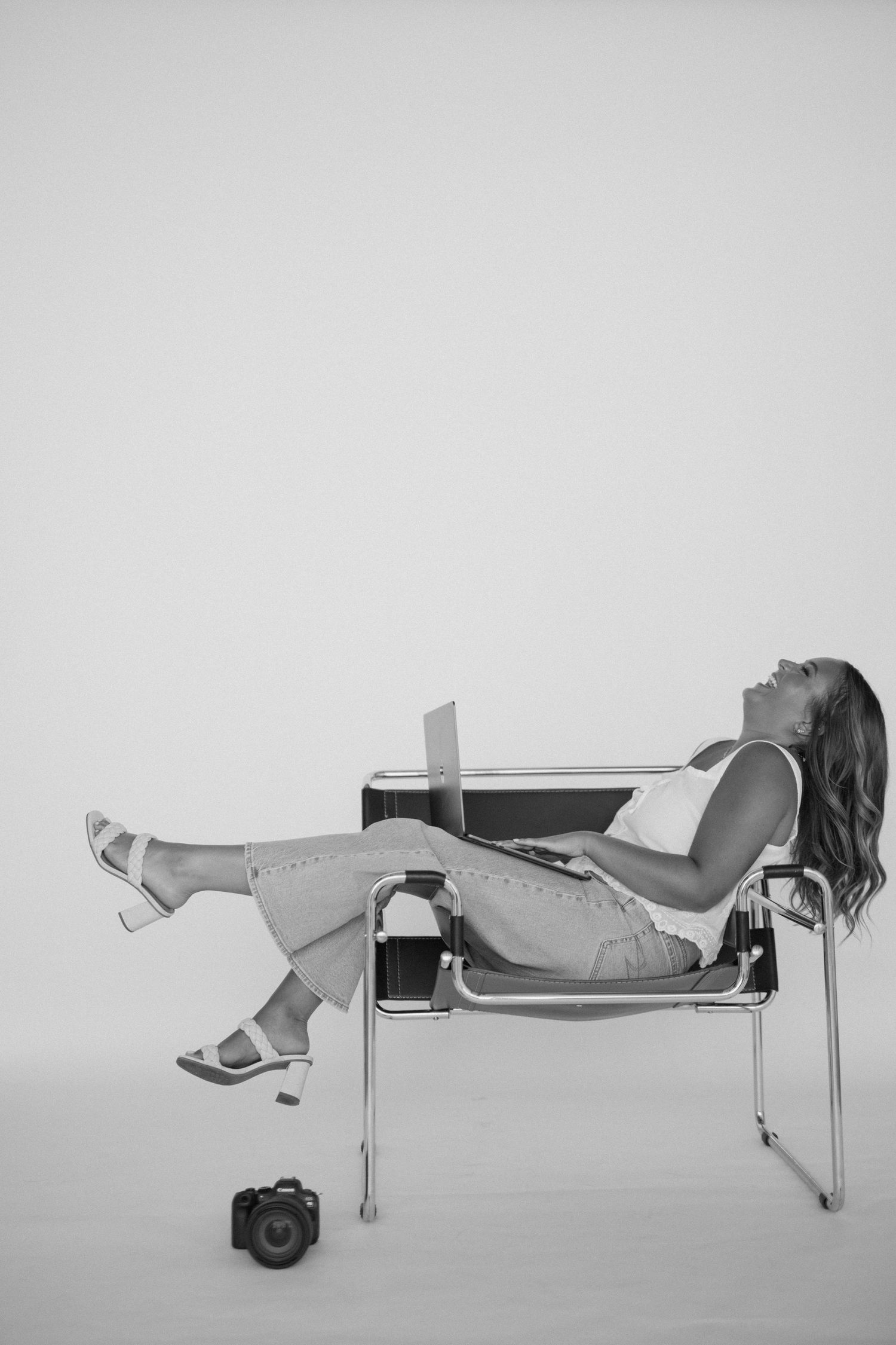 Black and white photo of a woman relaxing in a modern chair with a laptop, heels on, camera on the floor nearby.
