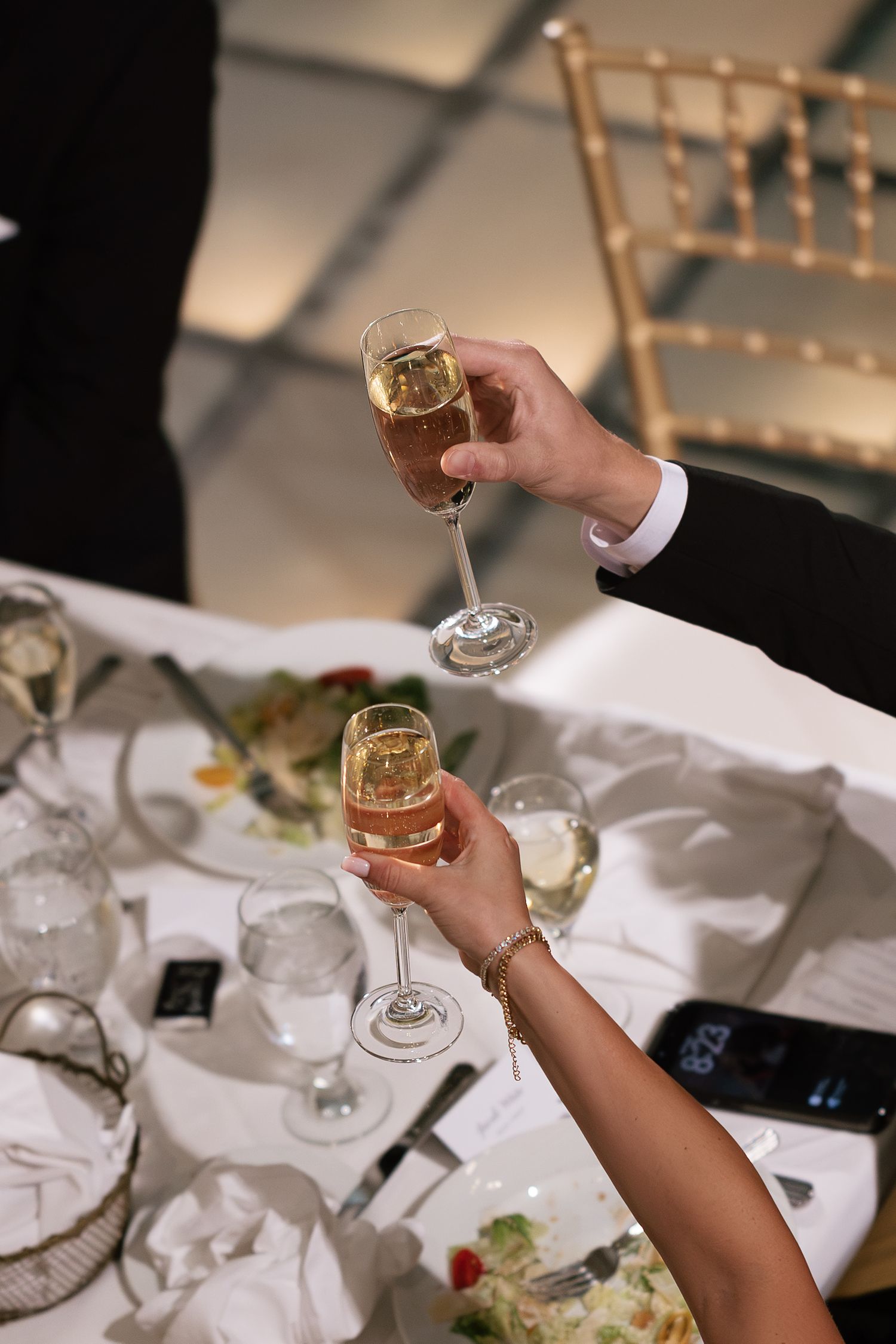 Two hands clinking champagne glasses at an elegant wedding reception dinner table with white linens.