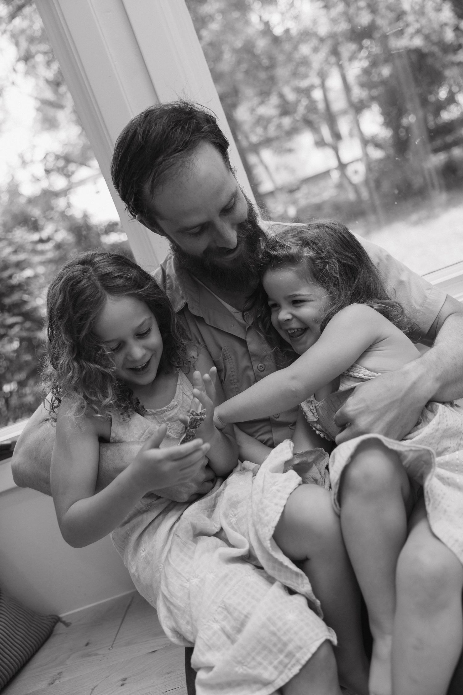 Black and white photo of bearded father laughing with two young daughters on a couch.