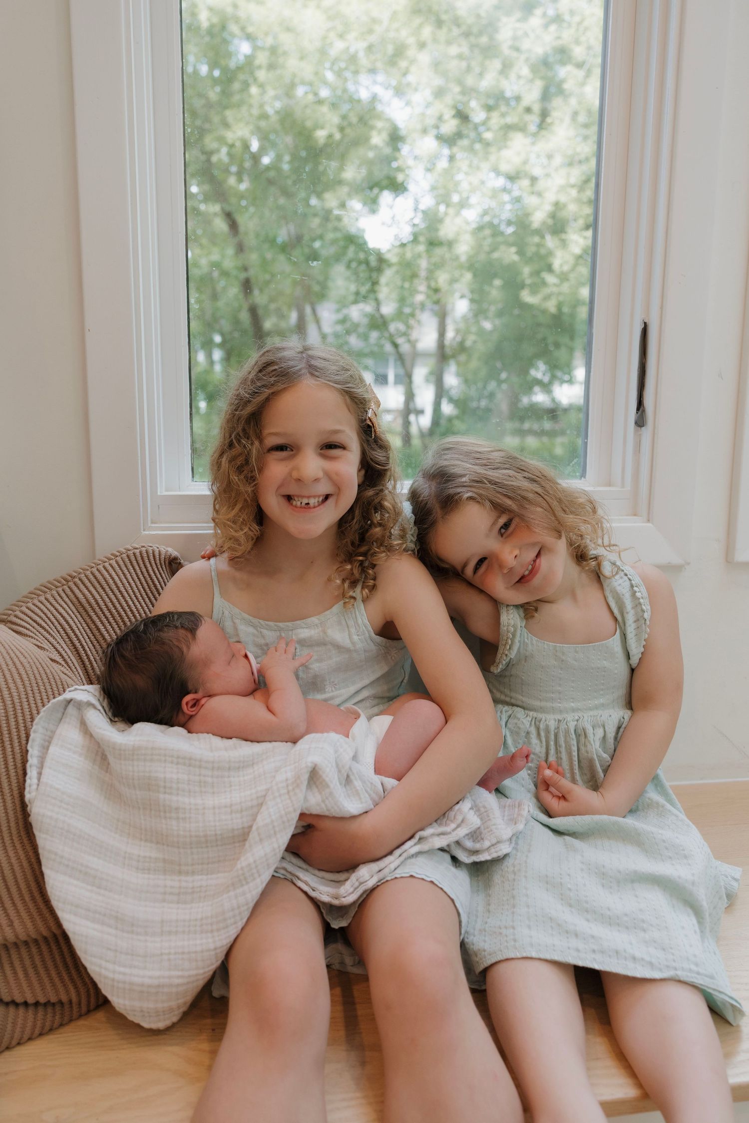 Two smiling sisters in mint dresses sitting together holding their newborn sibling indoors.