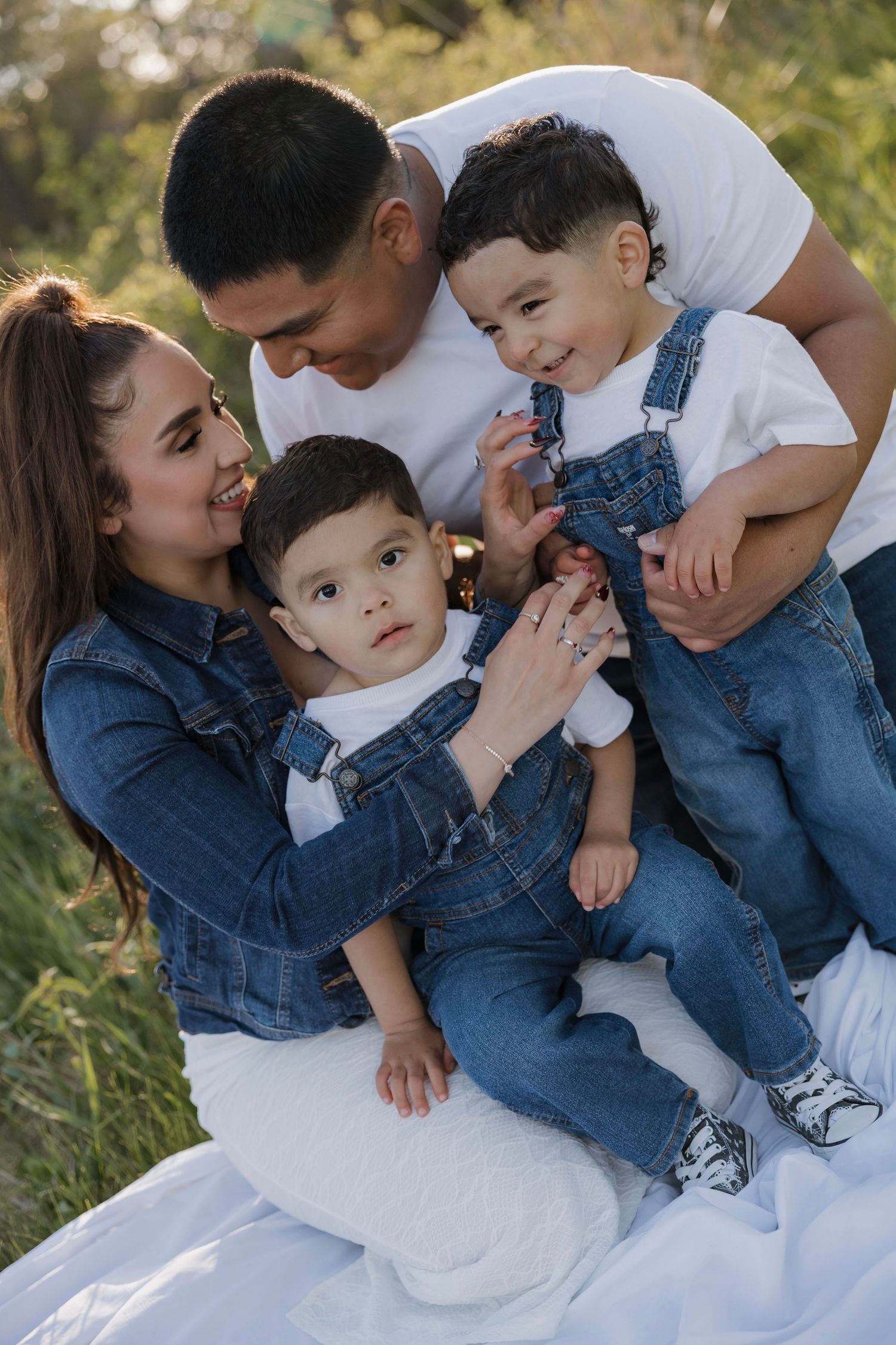 Happy young family with two toddler boys in matching denim overalls poses outdoors in a sunlit green field.