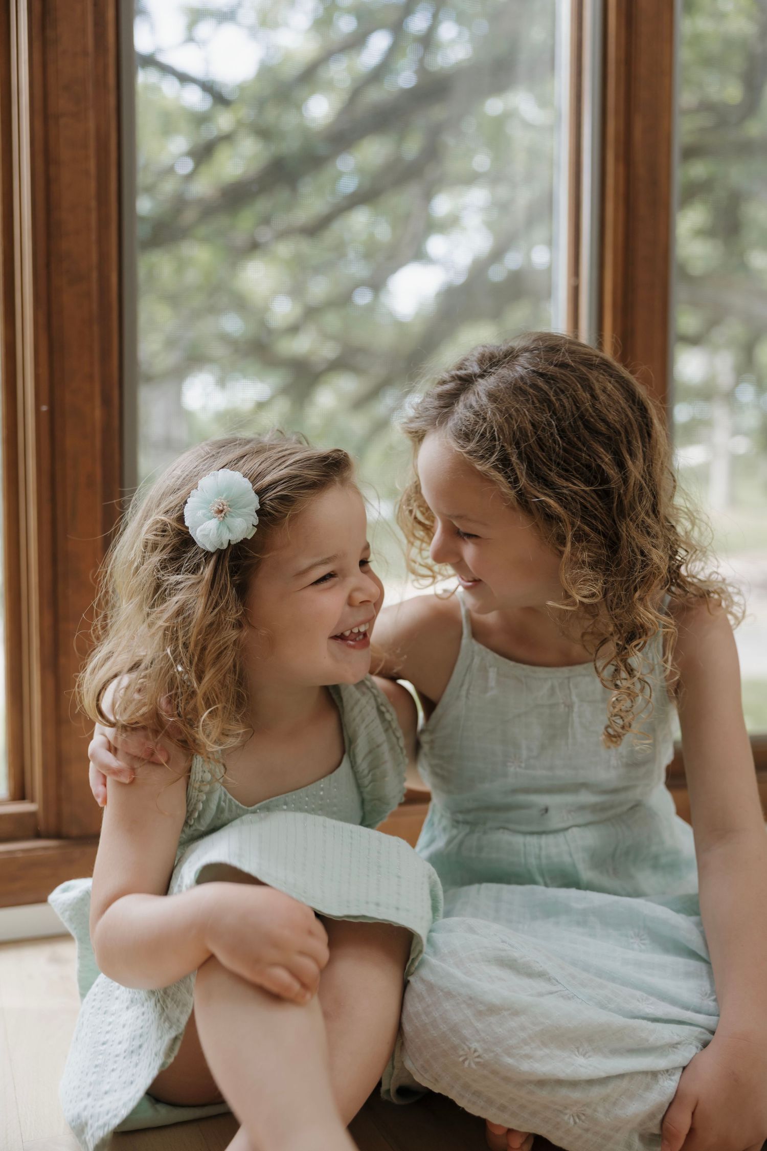 Two curly-haired sisters in matching mint dresses smiling at each other by a sunlit window.