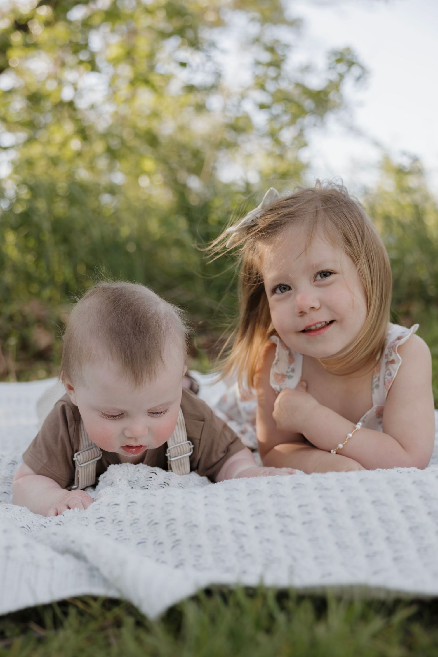 Toddler girl in floral dress smiles while lying on white blanket outdoors next to her baby sibling.