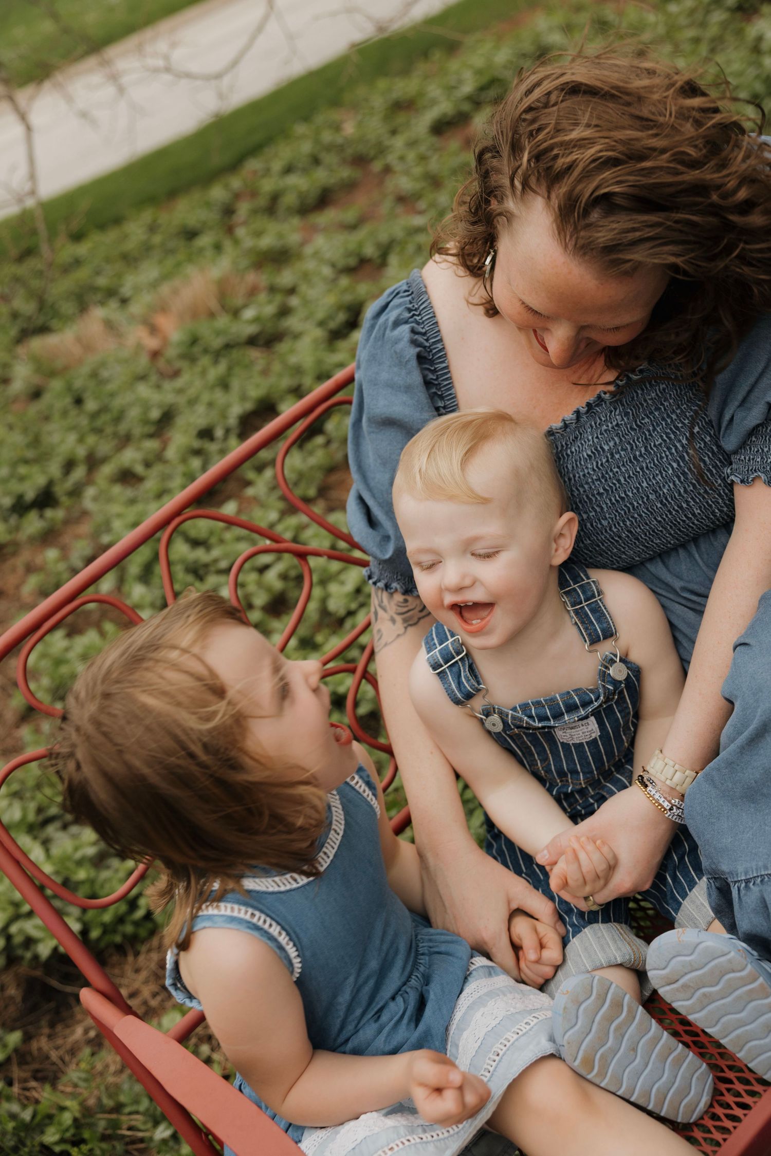 Mother sitting on red bench with two young children laughing outdoors.