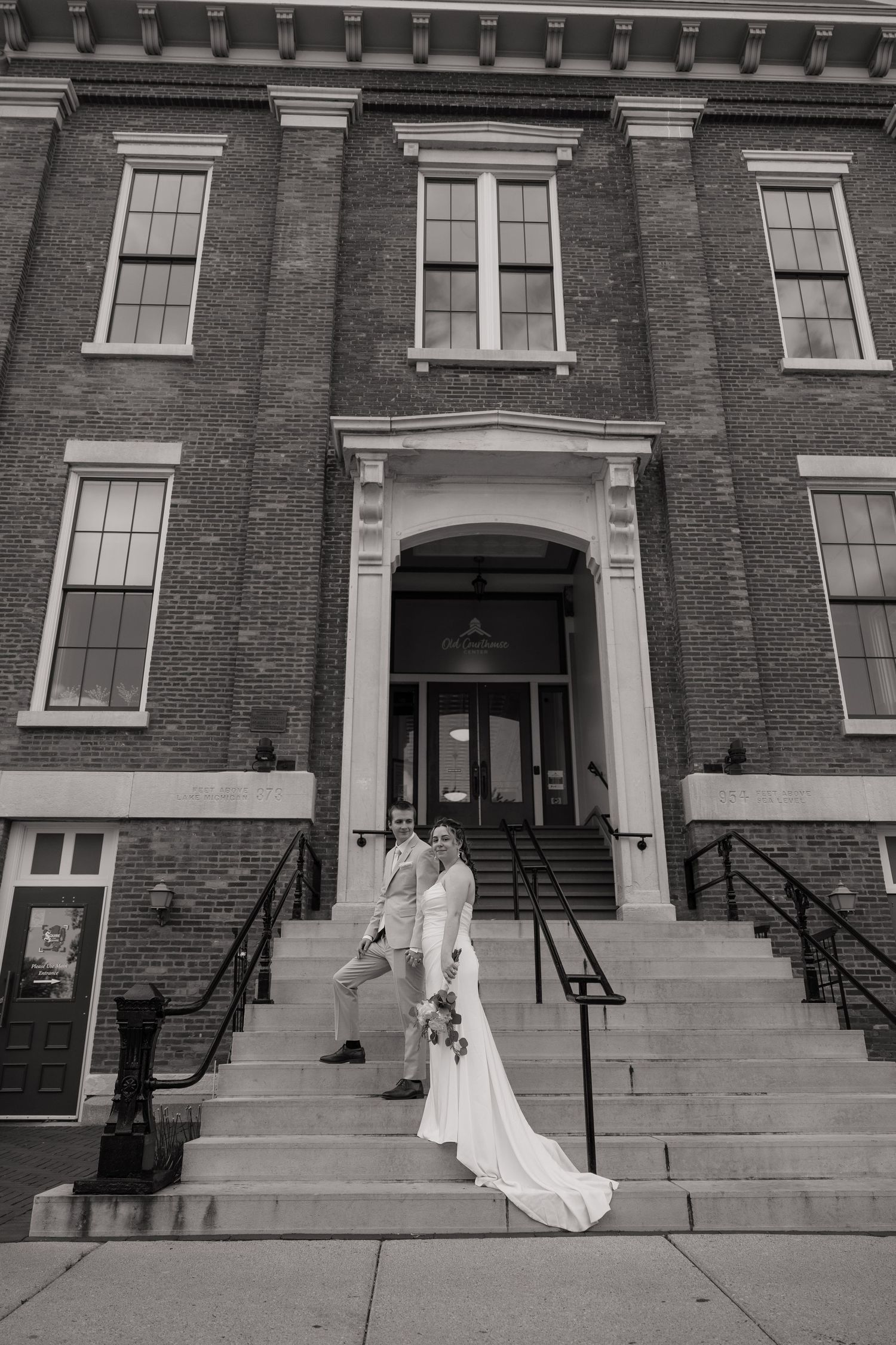 Black and white photo of a bride and groom posing on steps of a grand brick building on their wedding day.