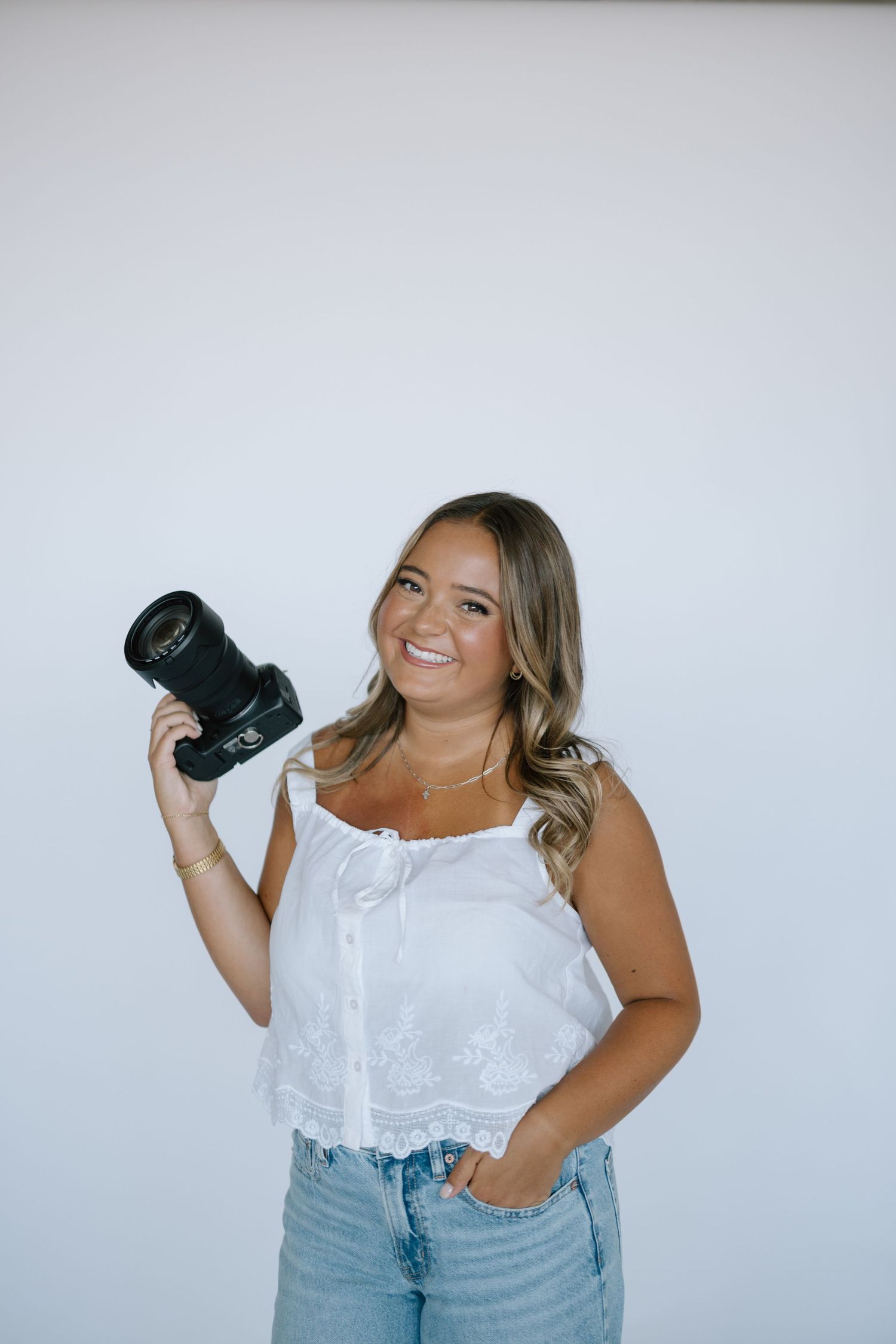 Smiling young woman holding a DSLR camera in a white crop top and jeans against a light background.